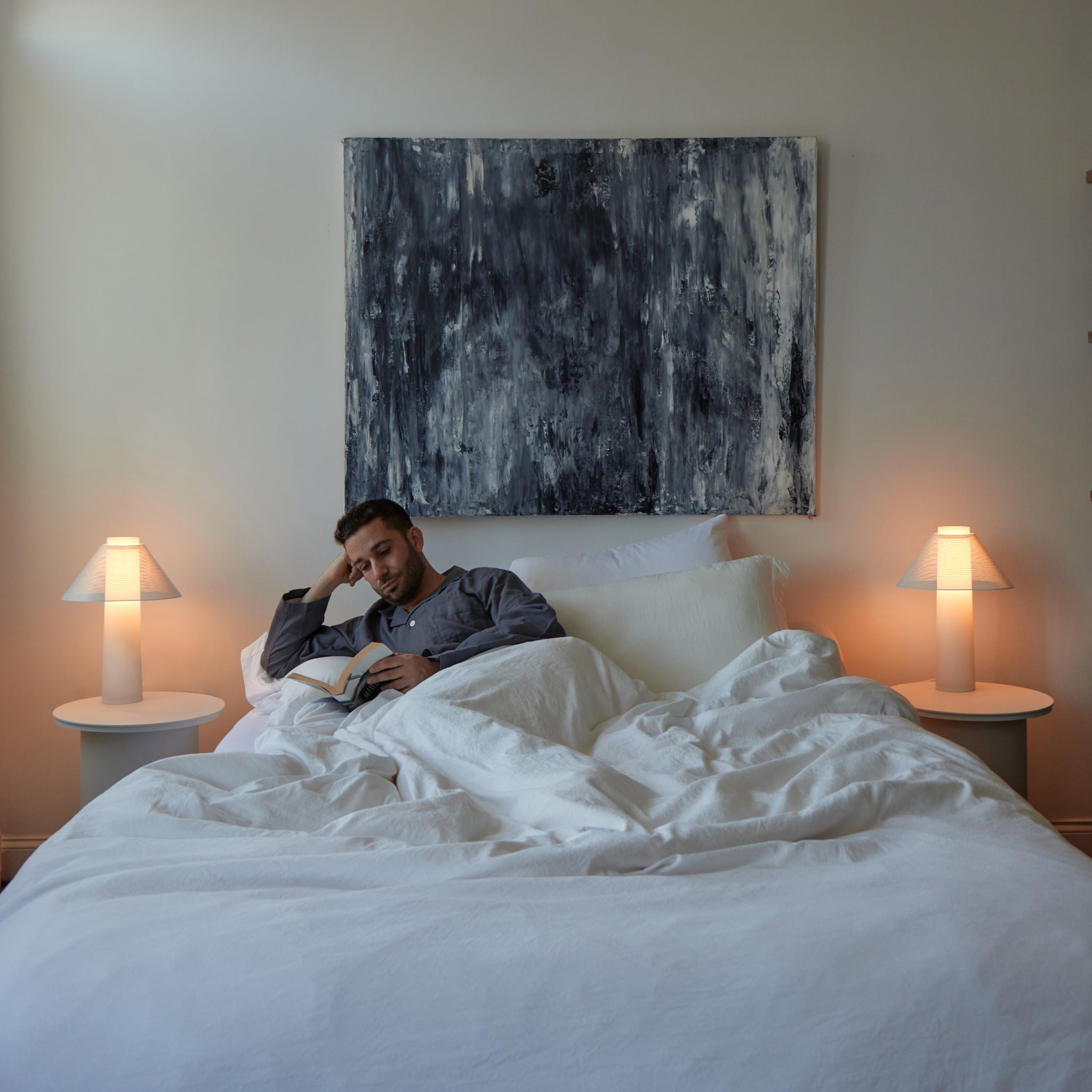Man lying in bed reading a book with a abstract painting on the wall behind him.