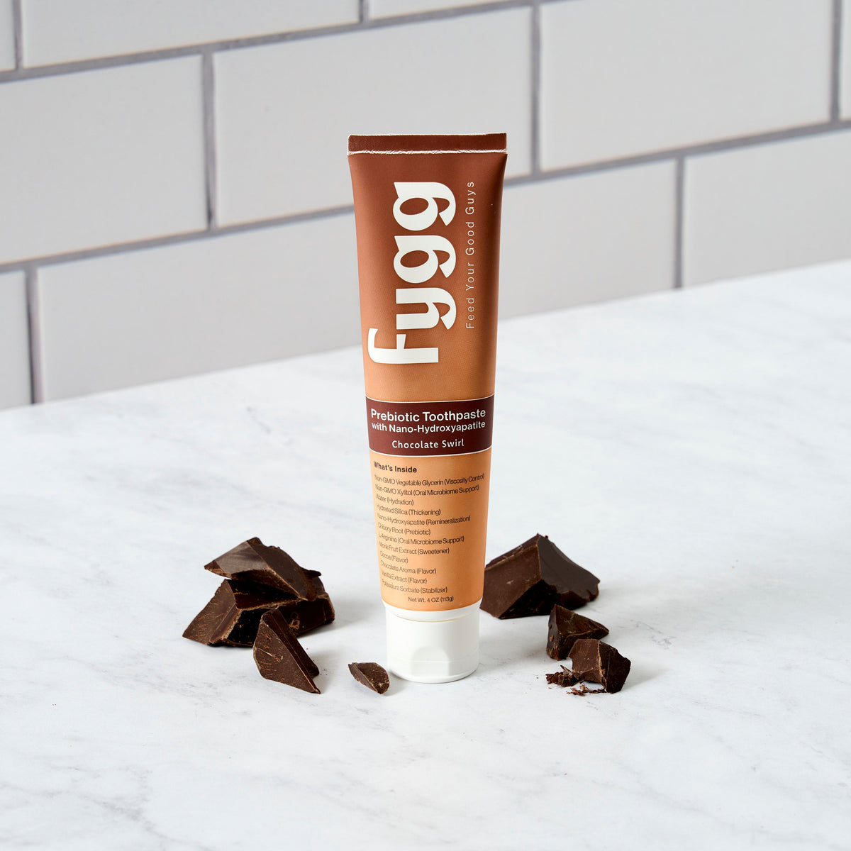 A tube of Fygg Prebiotic Toothpaste in Chocolate Sea Salt flavor, styled on a white marble surface with pieces of dark chocolate scattered around it, against a white subway tile background.