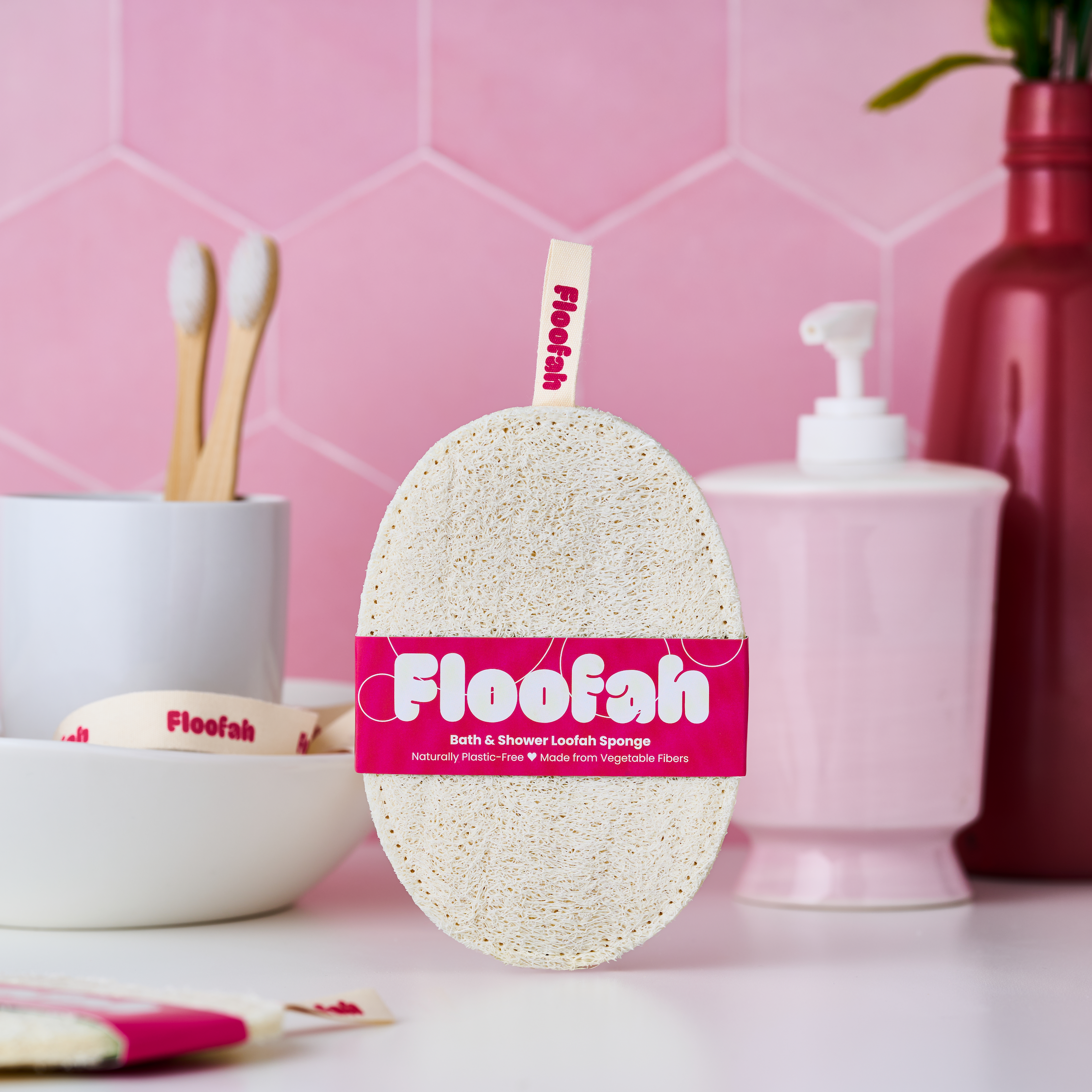 Floofah bath & shower loof sponge standing upright in a bathroom setting with pink hexagonal tiles, a pink soap dispenser, and white ceramic accessories in the background.