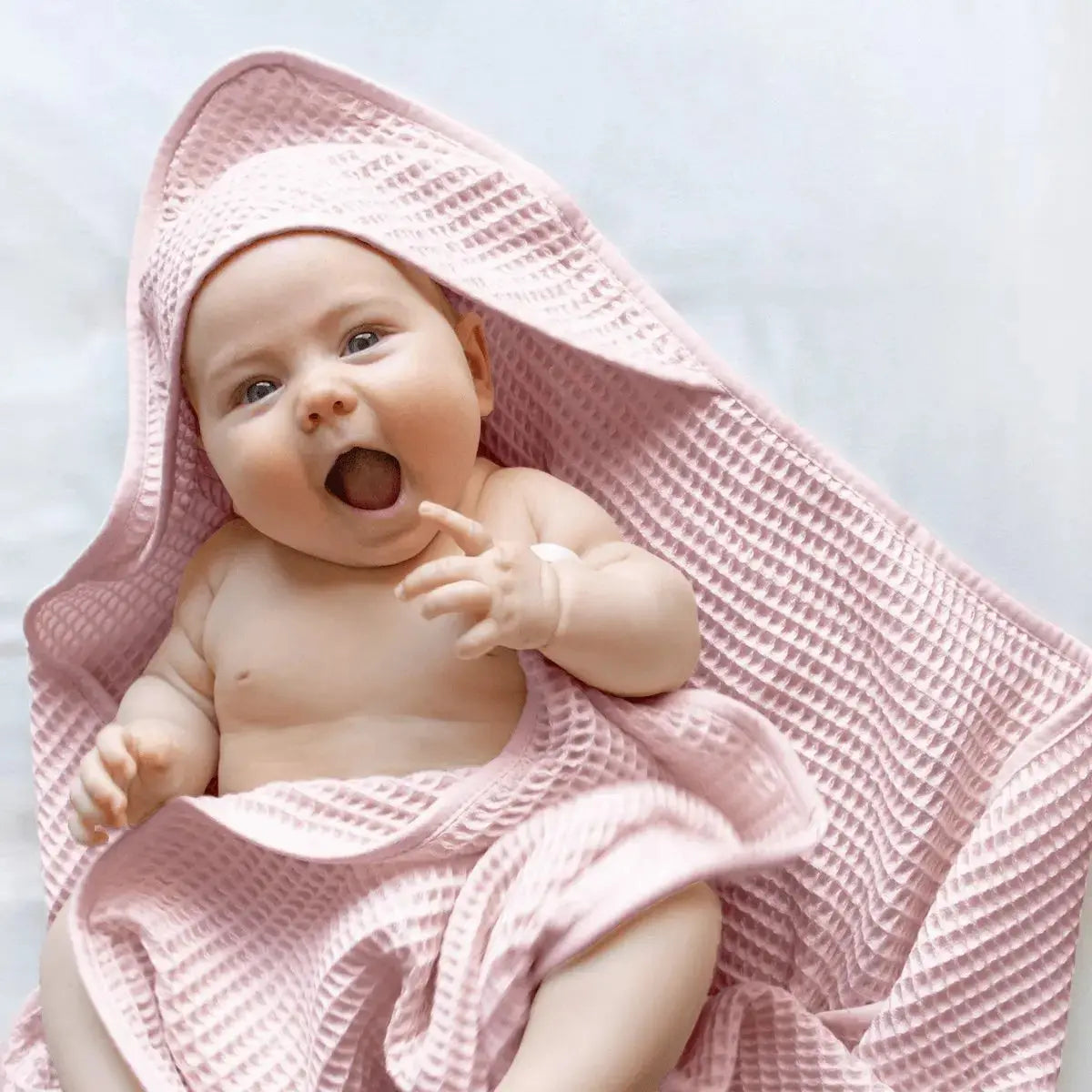 A baby wrapped in a pink waffle-weave hooded towel, looking surprised and smiling with their mouth open.