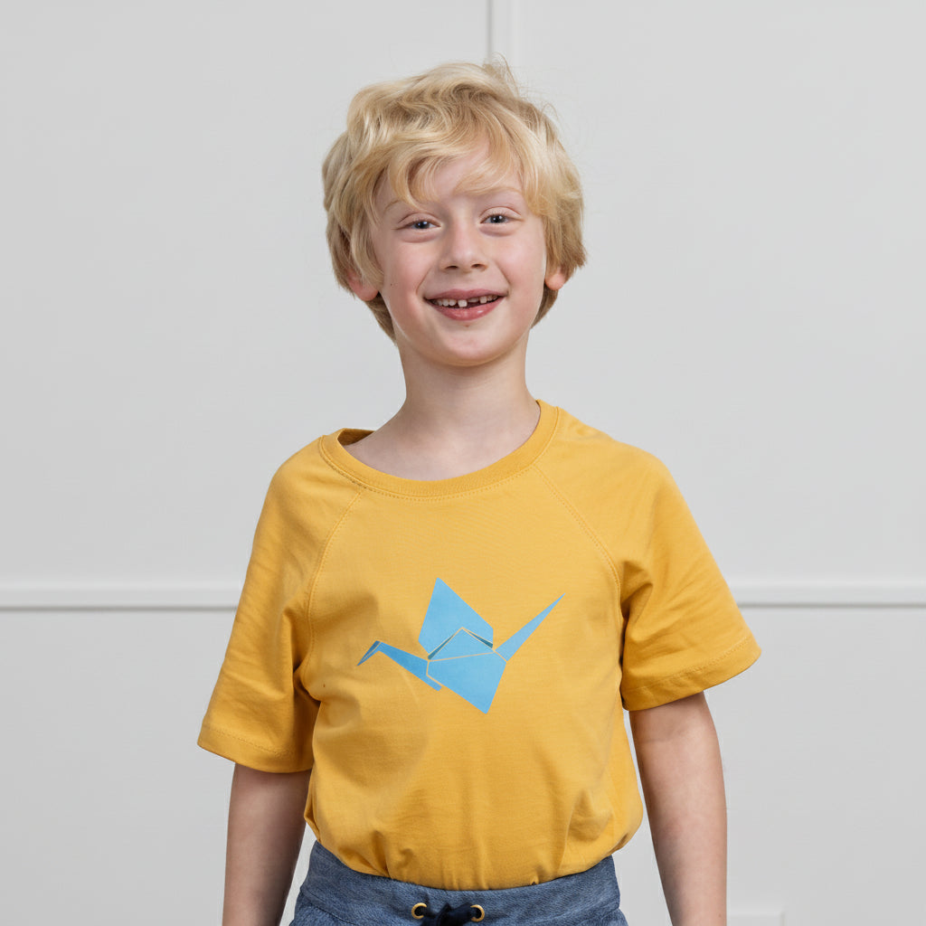 A young boy with blonde hair smiling, wearing a mustard yellow t-shirt with a light blue origami crane graphic on the front, set against a plain light gray wall.