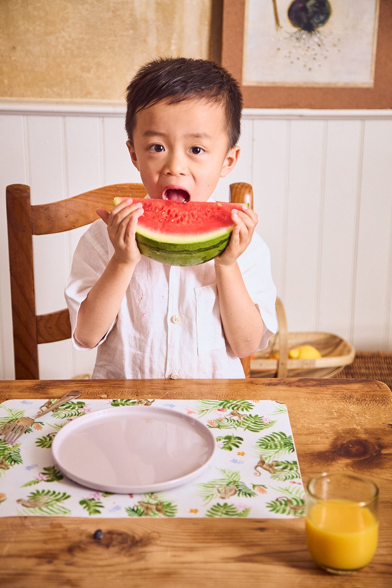 Child holding a watermelon slice at a dining table with a glass of orange juice.