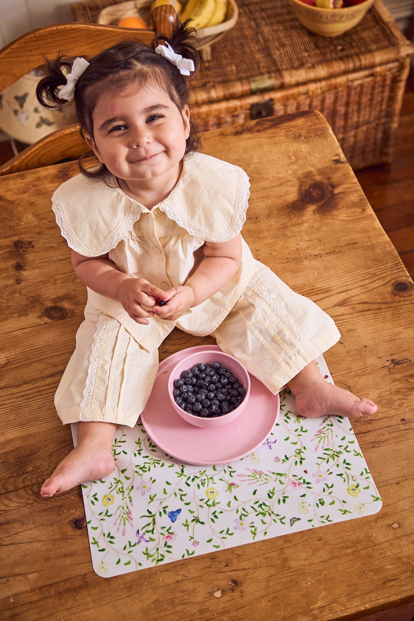 Child in a white dress sitting on a wooden table with a pink bowl of blueberries.