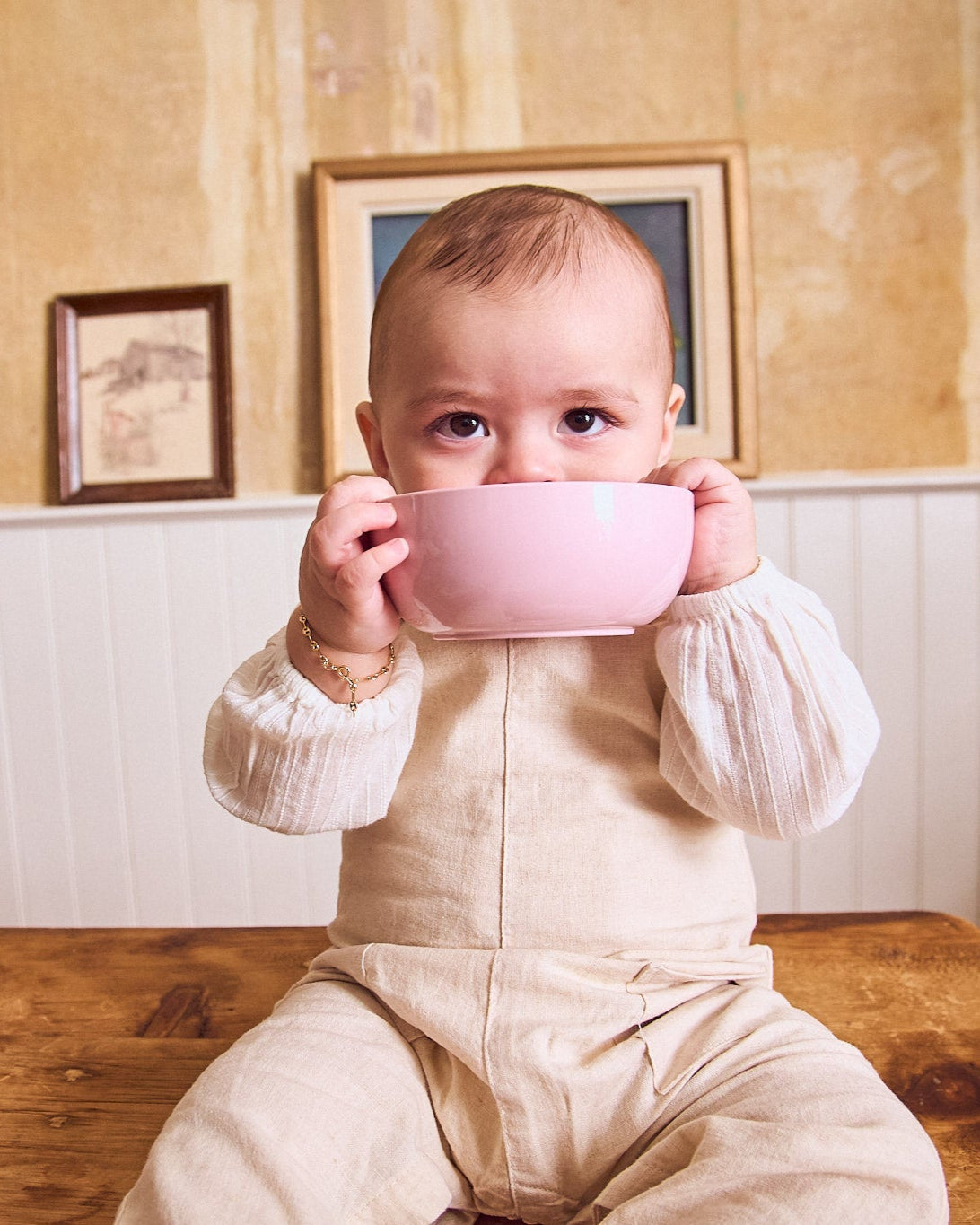 Baby holding a pink bowl in a room with framed pictures on the wall.