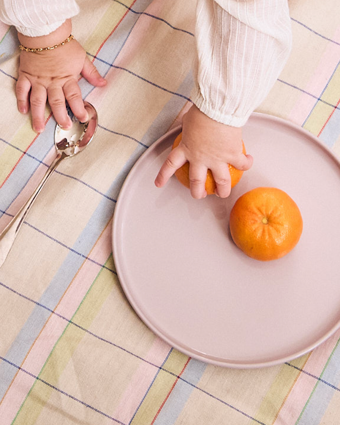 Child's hands with an orange on a pink plate on a checkered tablecloth