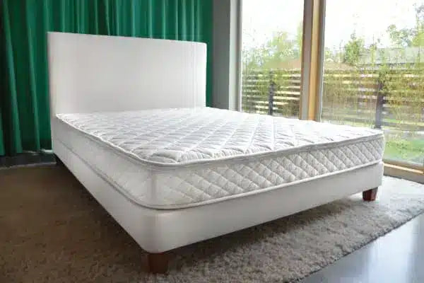 White mattress on a bed frame in a room with large windows and green curtains.