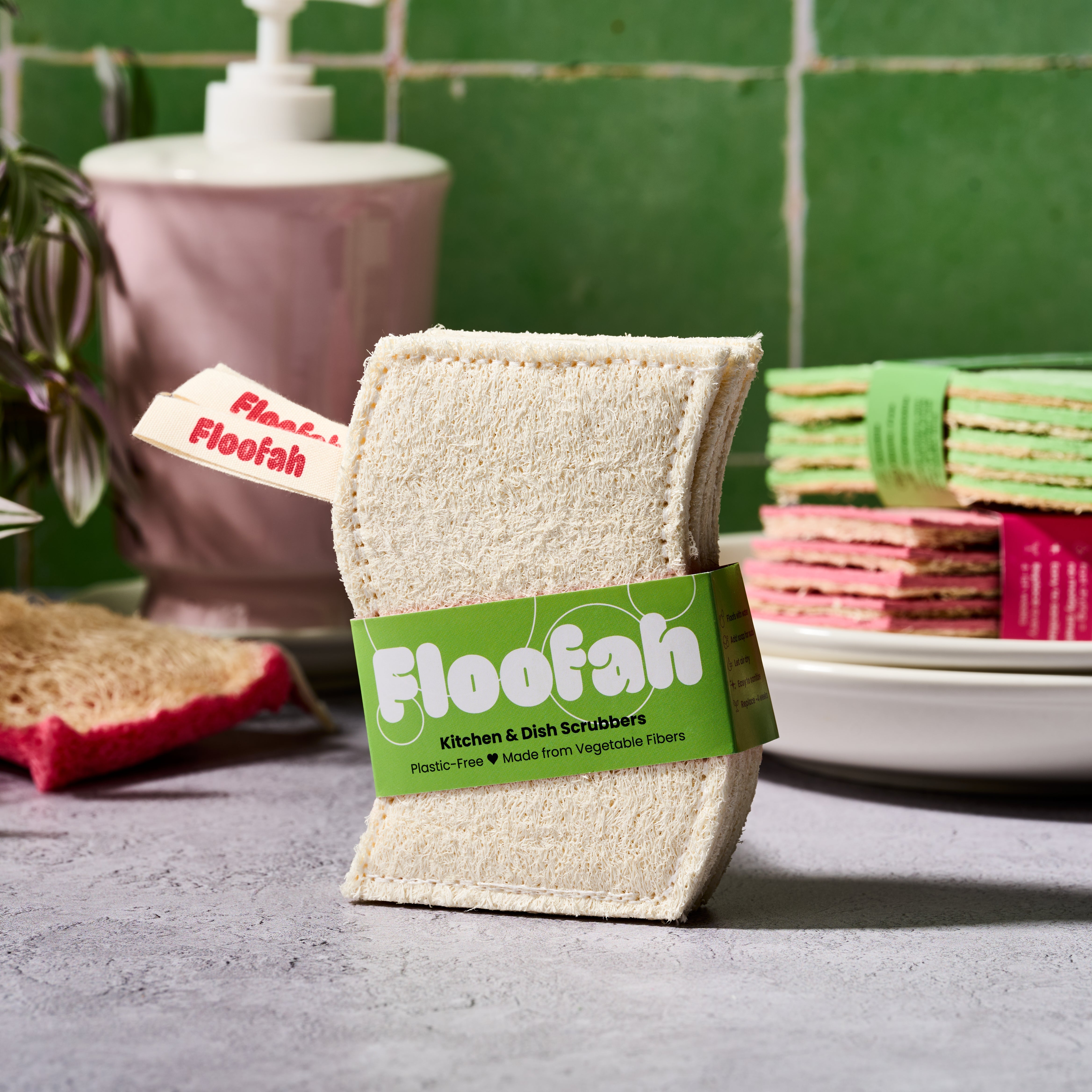A pack of Floofah Kitchen & Dish Scrubbers standing upright on a gray countertop, with a green label around the bundle. A pink soap dispenser, tiled green wall, and stacked scrubbers are blurred in the background.