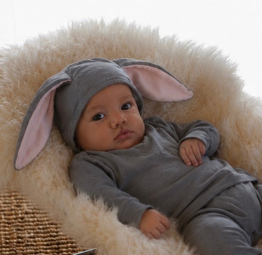 Baby wearing a gray bunny costume with large ears, lying on a fluffy white surface.