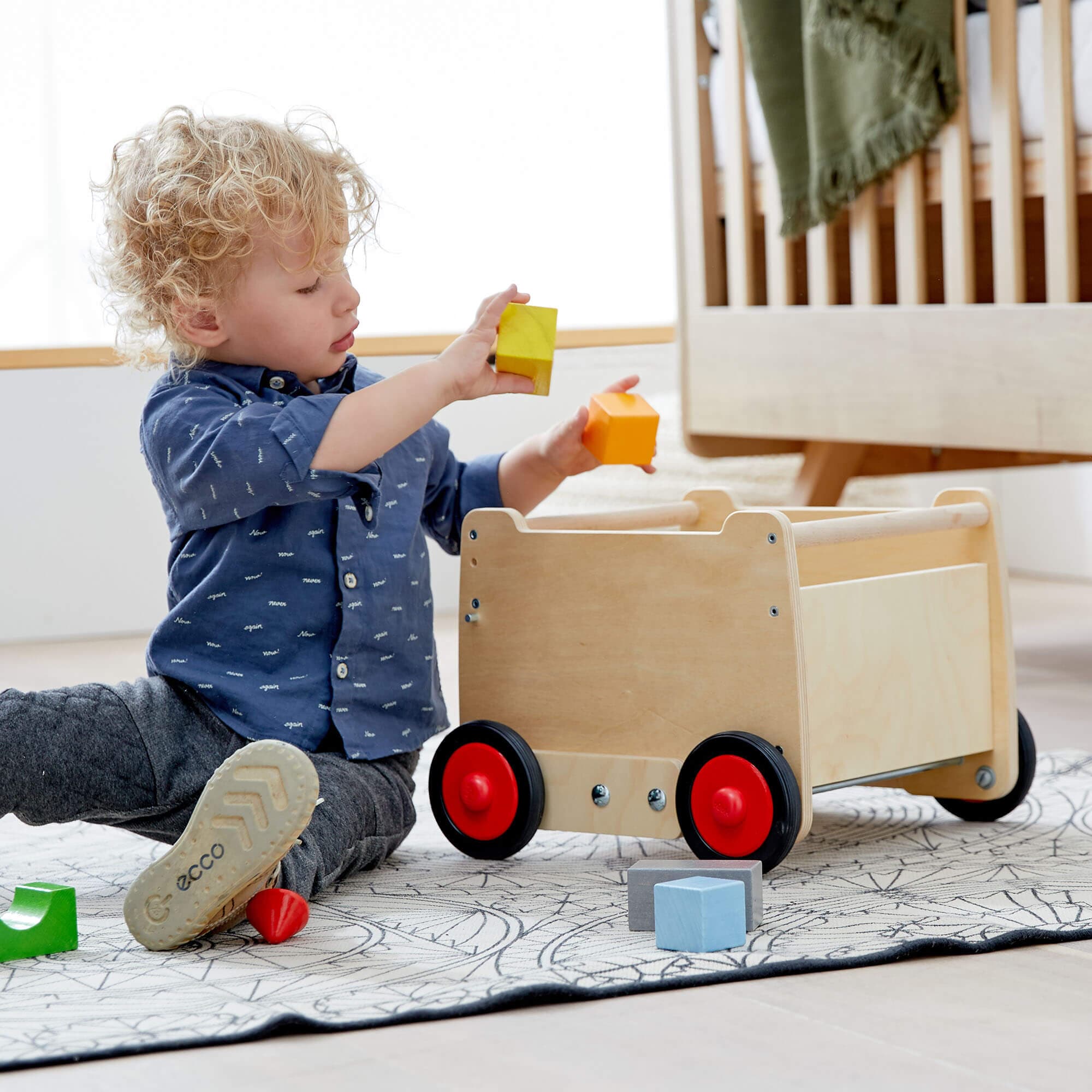 Child playing with colorful blocks next to a wooden cart on a rug.