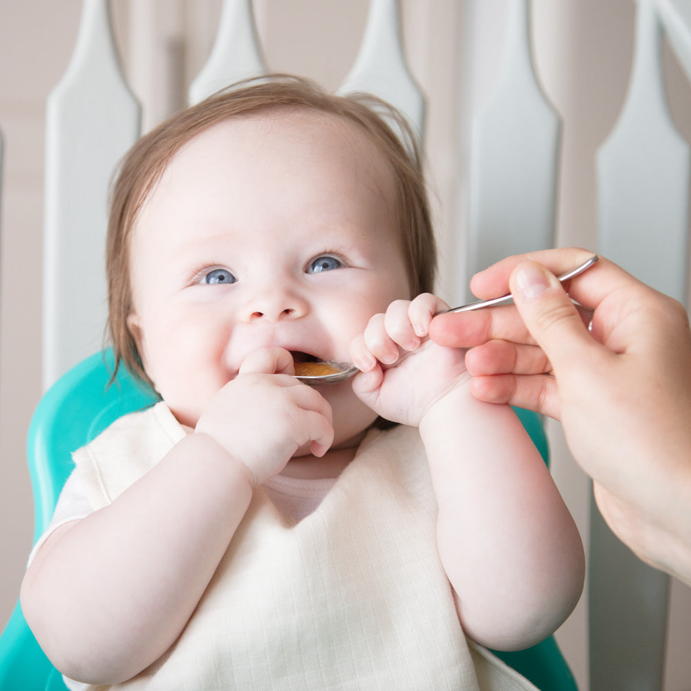 Baby being fed with a spoon by an adult, sitting in a high chair.