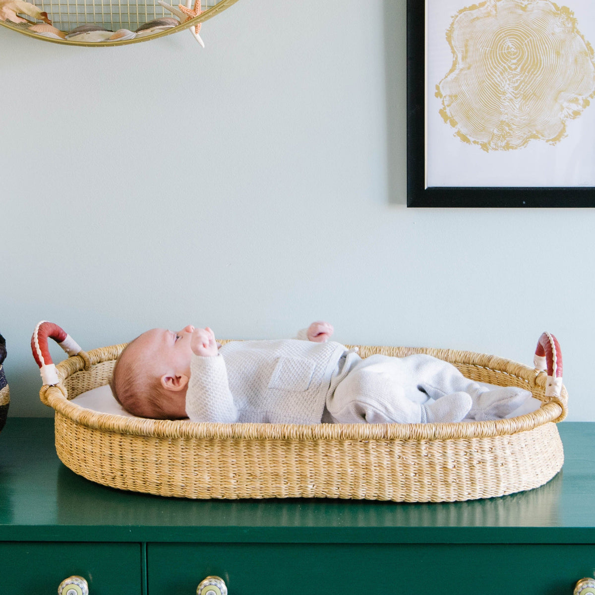 Baby lying in a woven bassinet against a light blue wall with decorative elements.