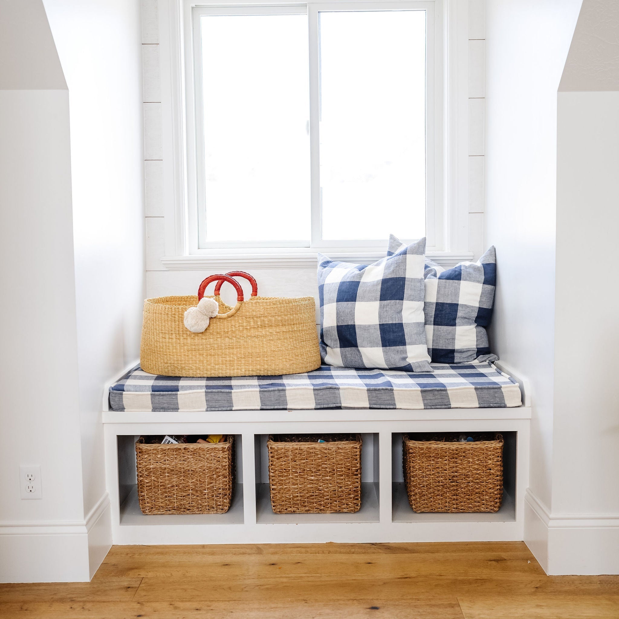 Bench with plaid cushion and storage baskets against a white wall with a window.