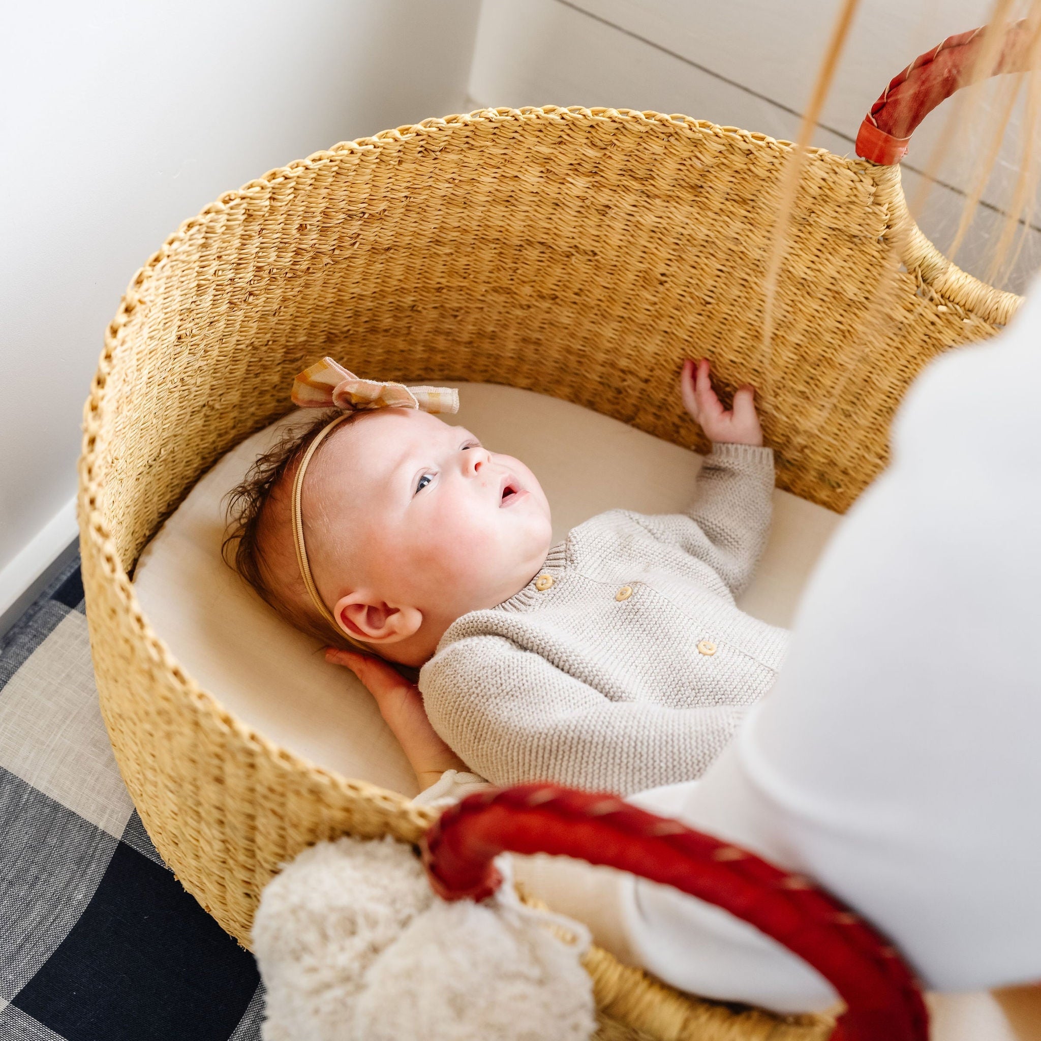 Baby lying in a woven crib with a soft toy, wearing a bow headband.