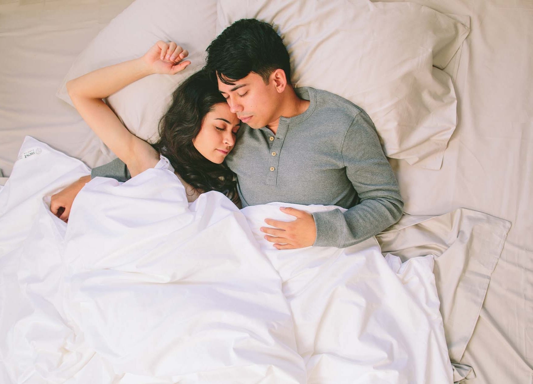 Couple lying in bed together, embracing under white sheets.