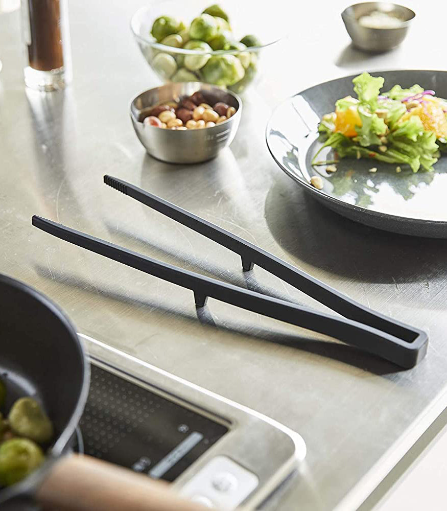 Black kitchen tongs on a kitchen counter with bowls of vegetables and a plate of salad.
