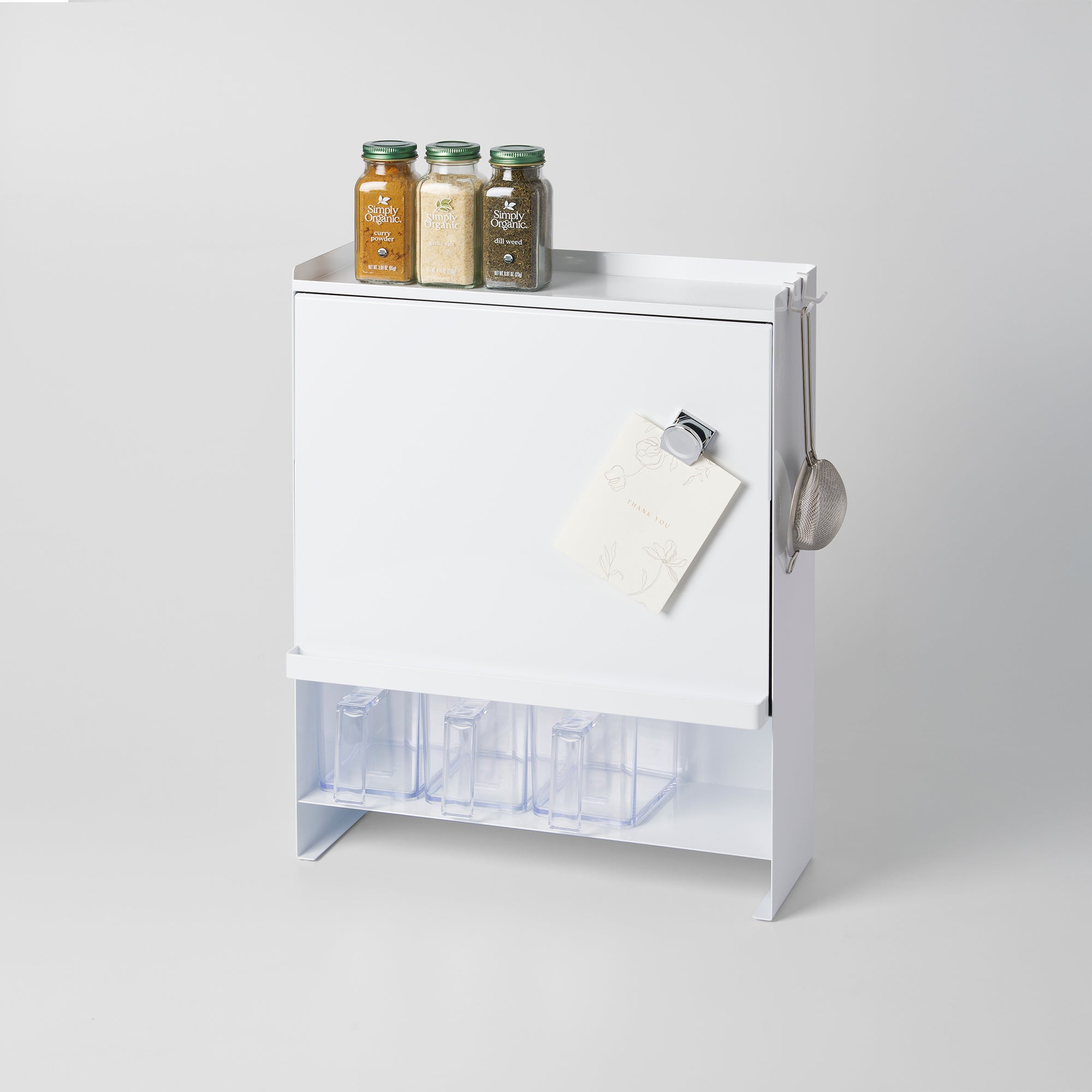 White kitchen cabinet with spice jars on top and a magnetic whiteboard attached, on a light gray background.