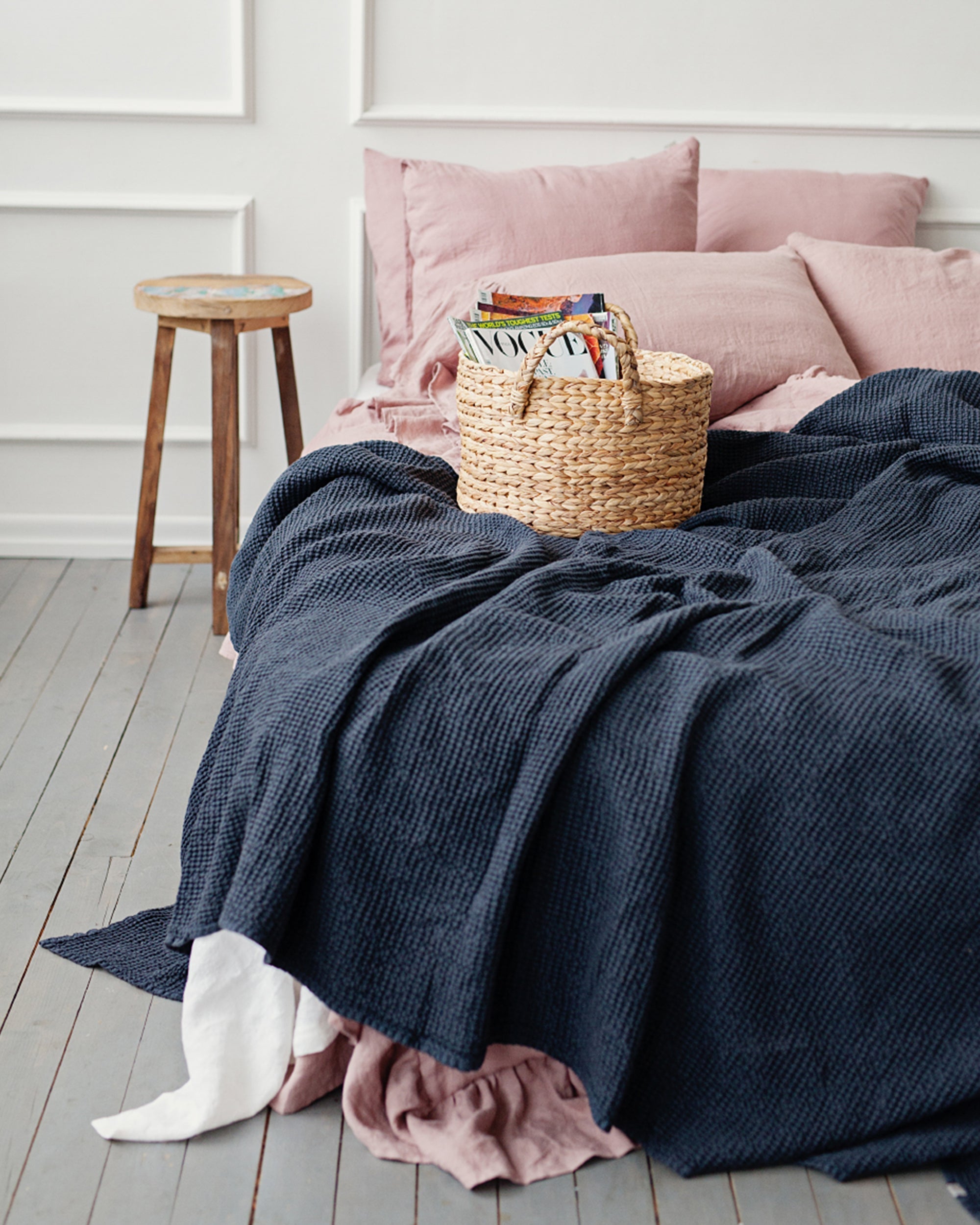 Bed with pink pillows, blue blanket, and a basket of books on a wooden floor.