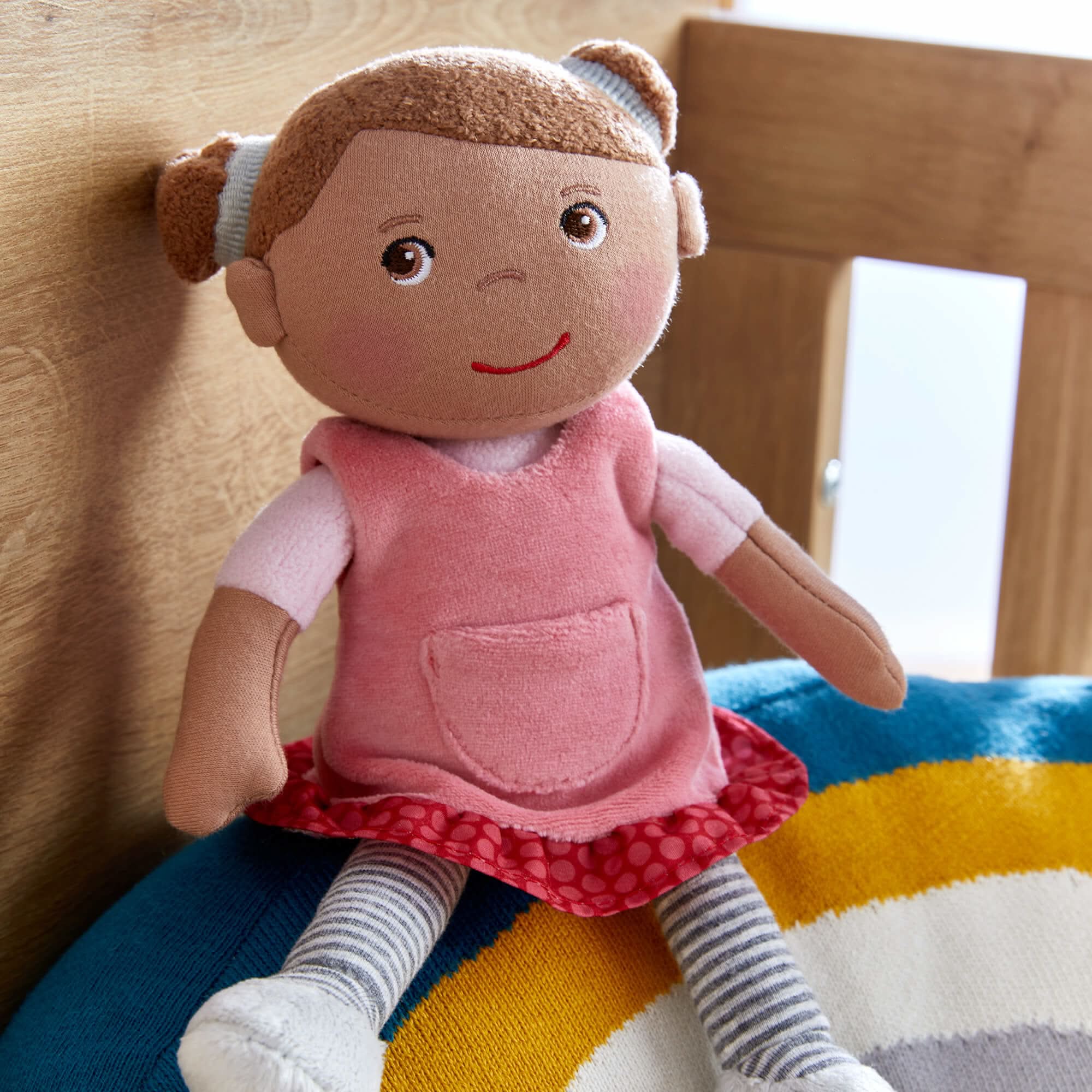 Plush doll with a pink dress sitting on a colorful striped blanket.