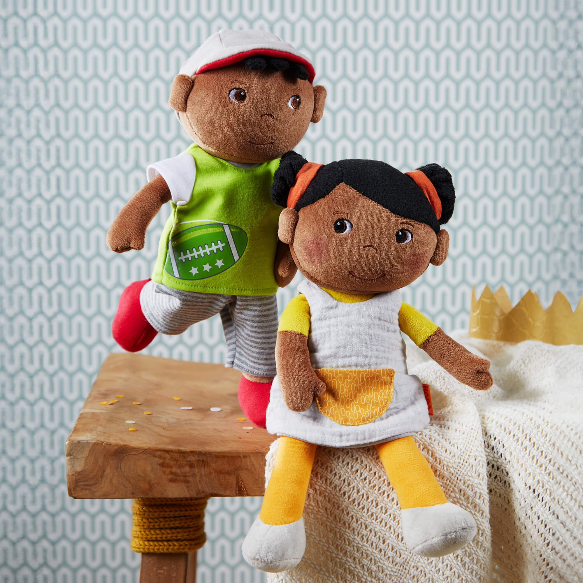 Two dolls, one in sports attire and the other in a dress, sitting on a wooden stool against a patterned wall.