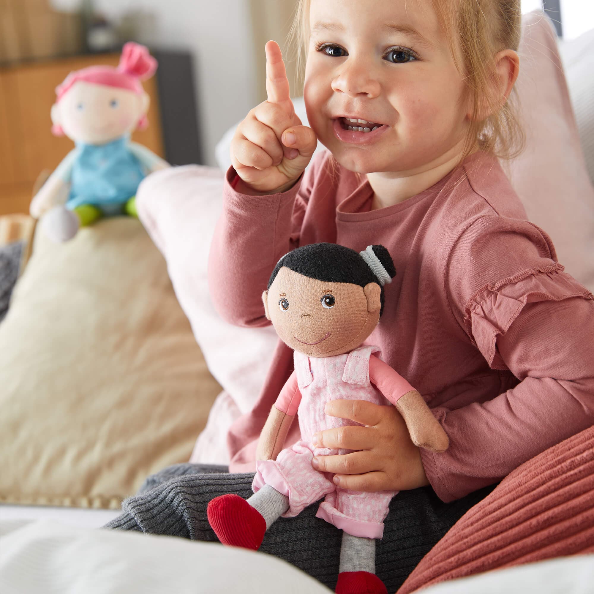 Child holding a doll and pointing upwards with a smile, surrounded by soft furnishings.