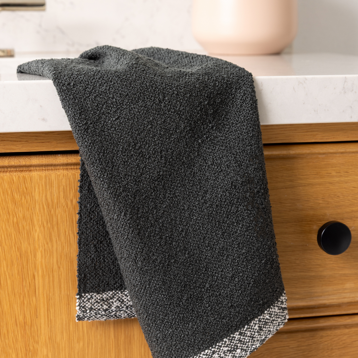 A dark charcoal gray textured kitchen towel with a black and white patterned border, draped over a wooden kitchen drawer next to a white marble countertop.