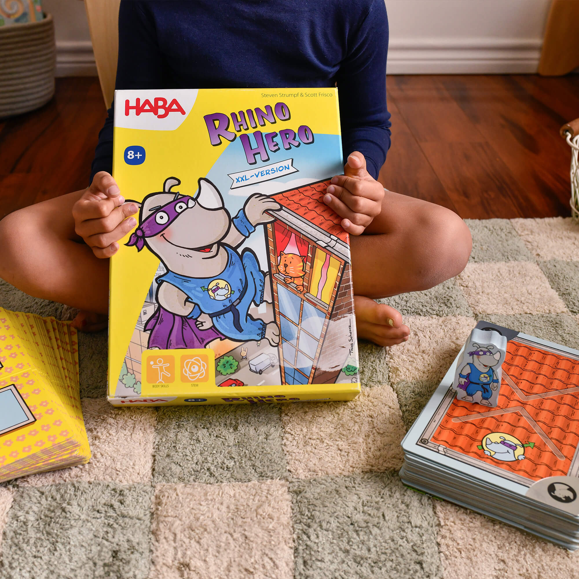 Child holding a HABA Rhino Hero game box on a carpeted floor with game cards nearby.