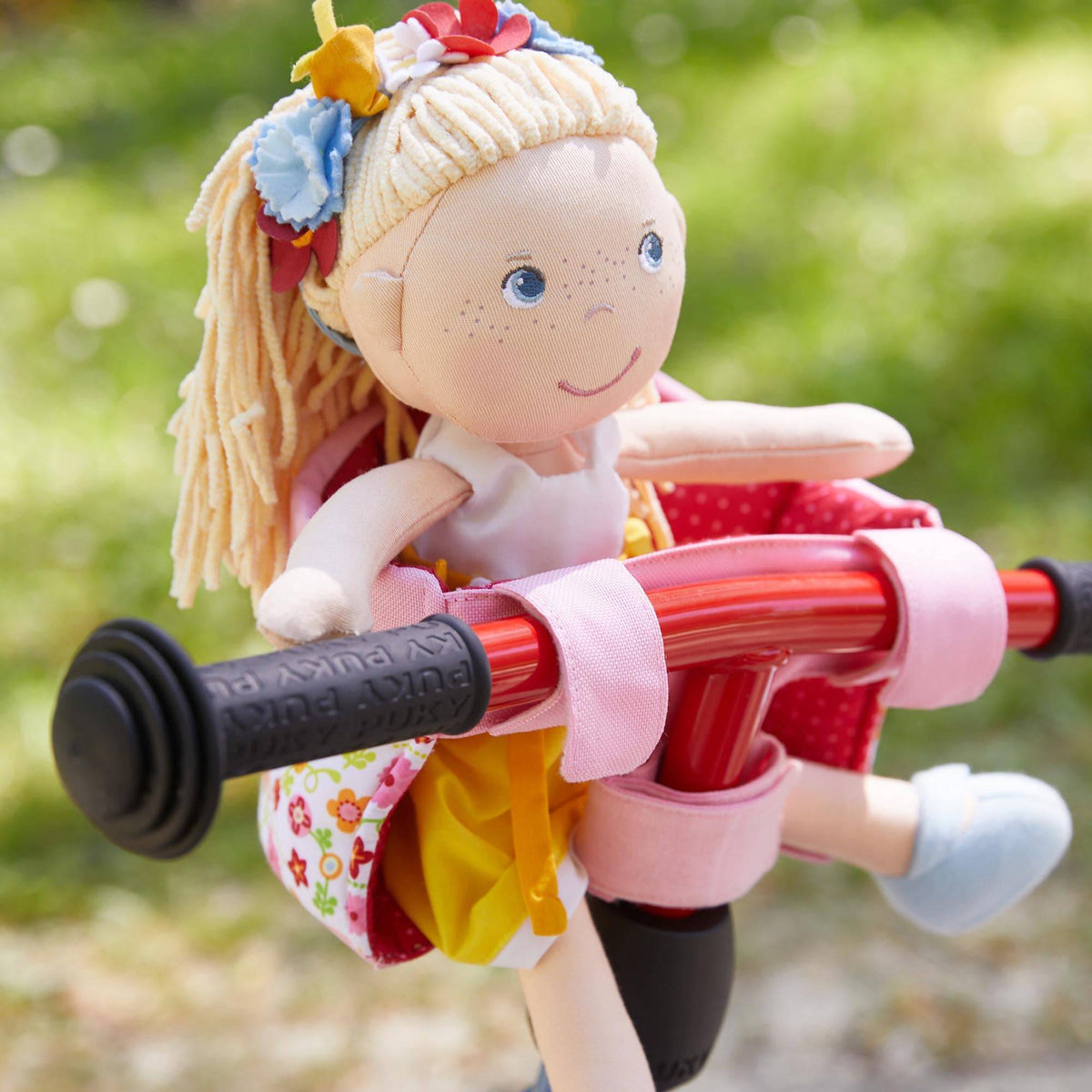Doll with blonde hair and floral headband sitting on a toy scooter with a blurred green background