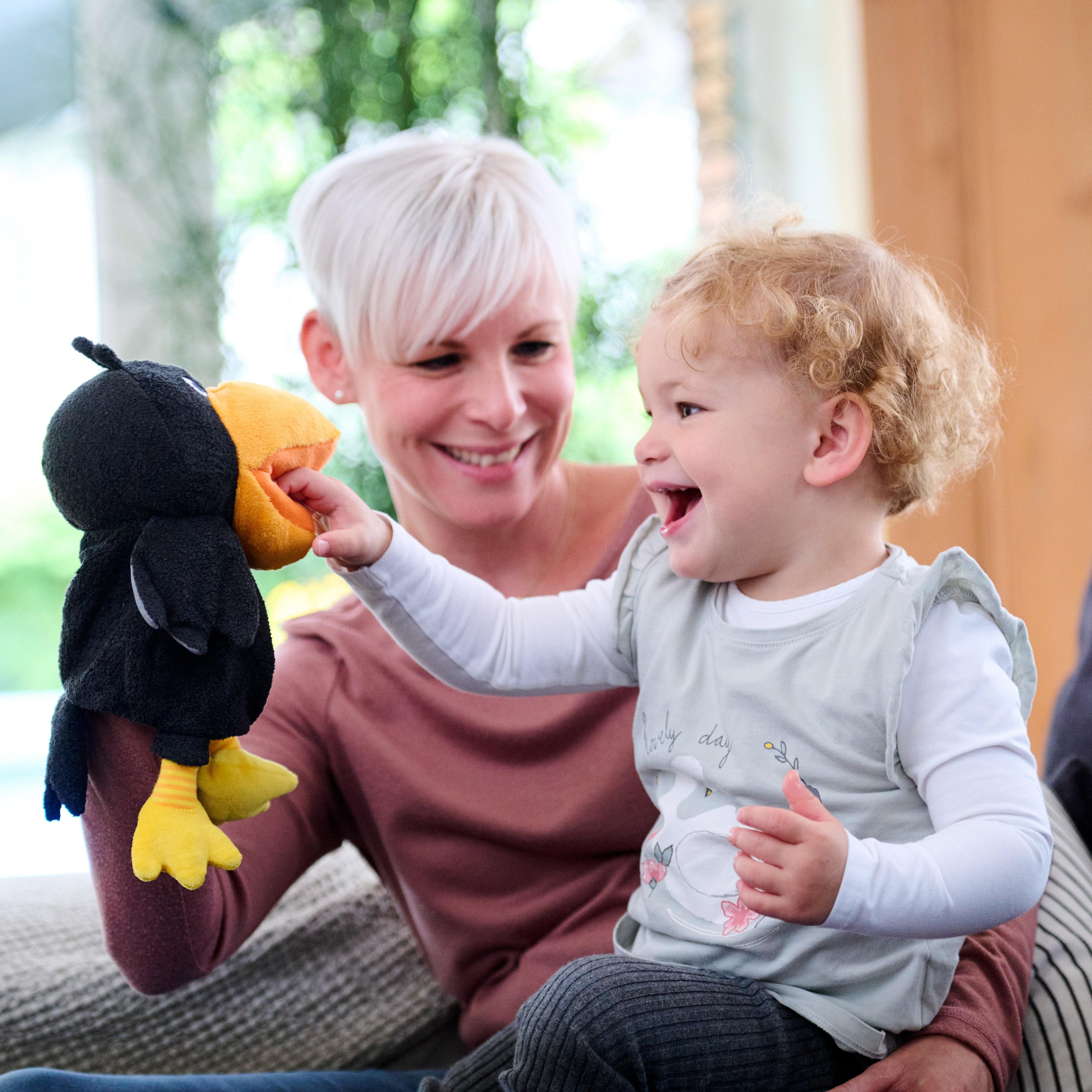 Woman holding a child with a toy, both smiling indoors