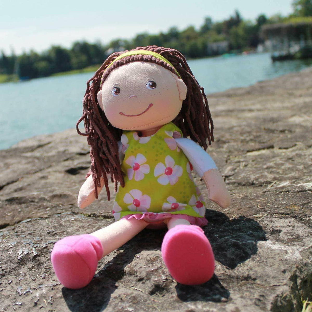 Plush doll with brown hair and a green dress with pink flowers sitting on a stone surface by a body of water.