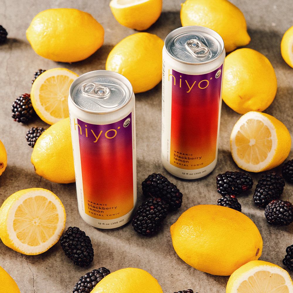 Two cans of 'Hiyo' blackberry lemonade surrounded by lemons and blackberries on a textured surface.