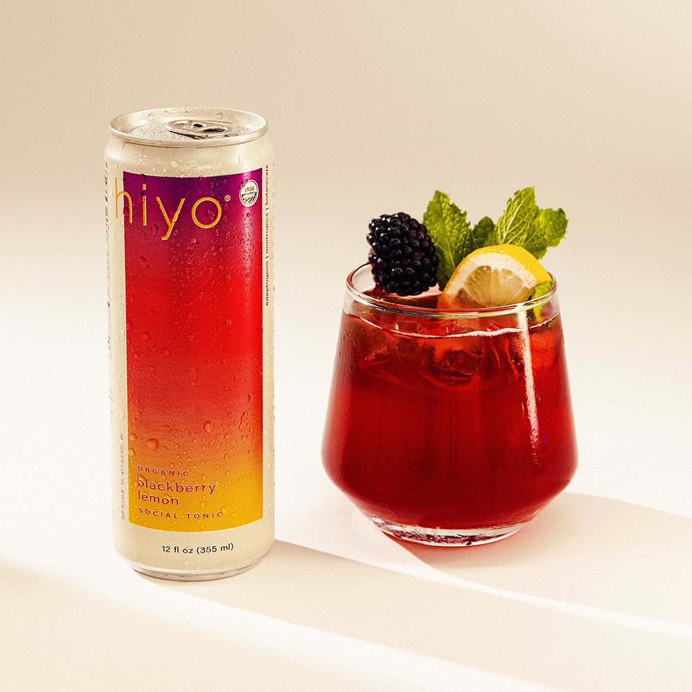 Can of hiyo blackberry lemon soda next to a glass of the same drink with garnishes on a light background