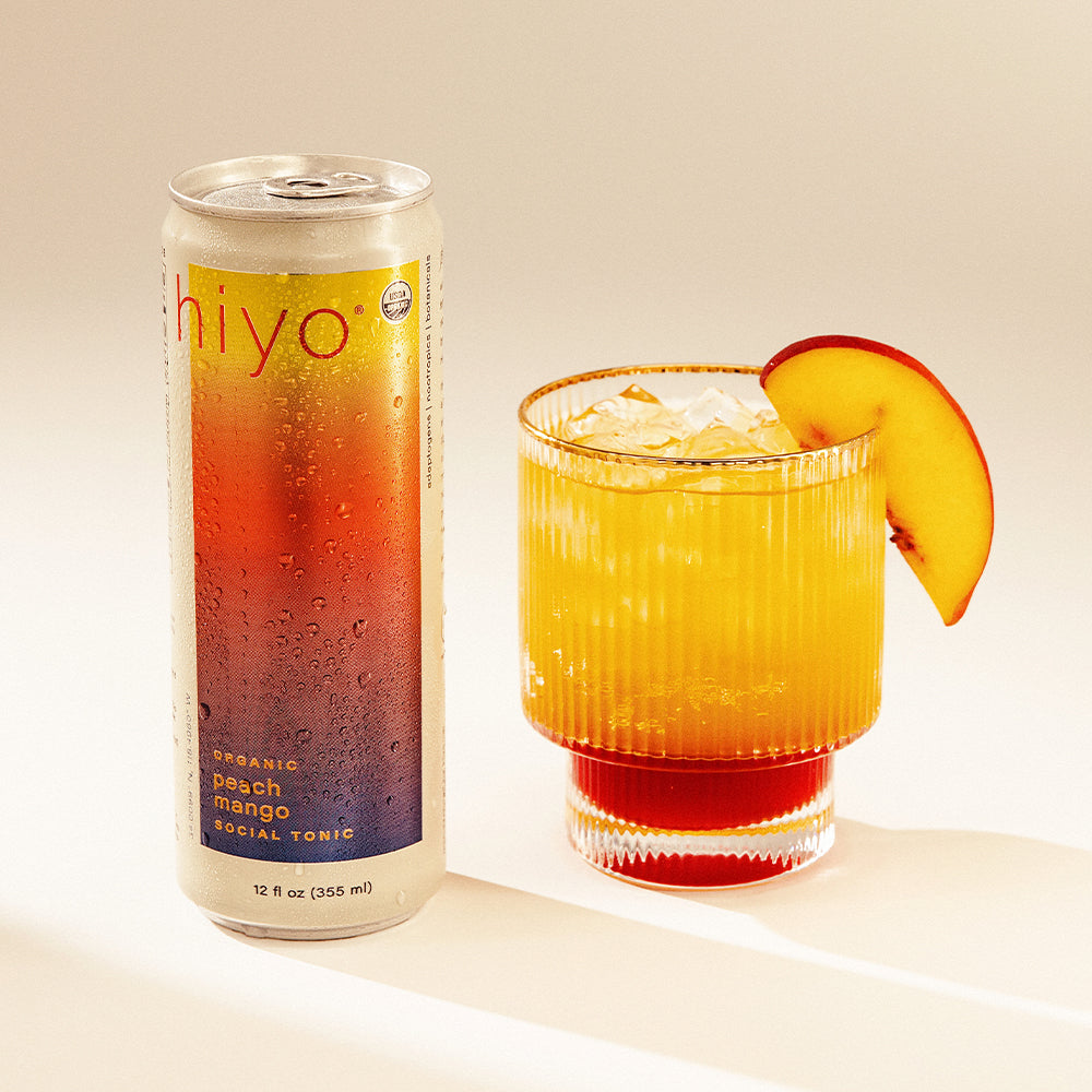 Can of hiyo peach mango social tonic next to a glass with a peach slice on a beige background