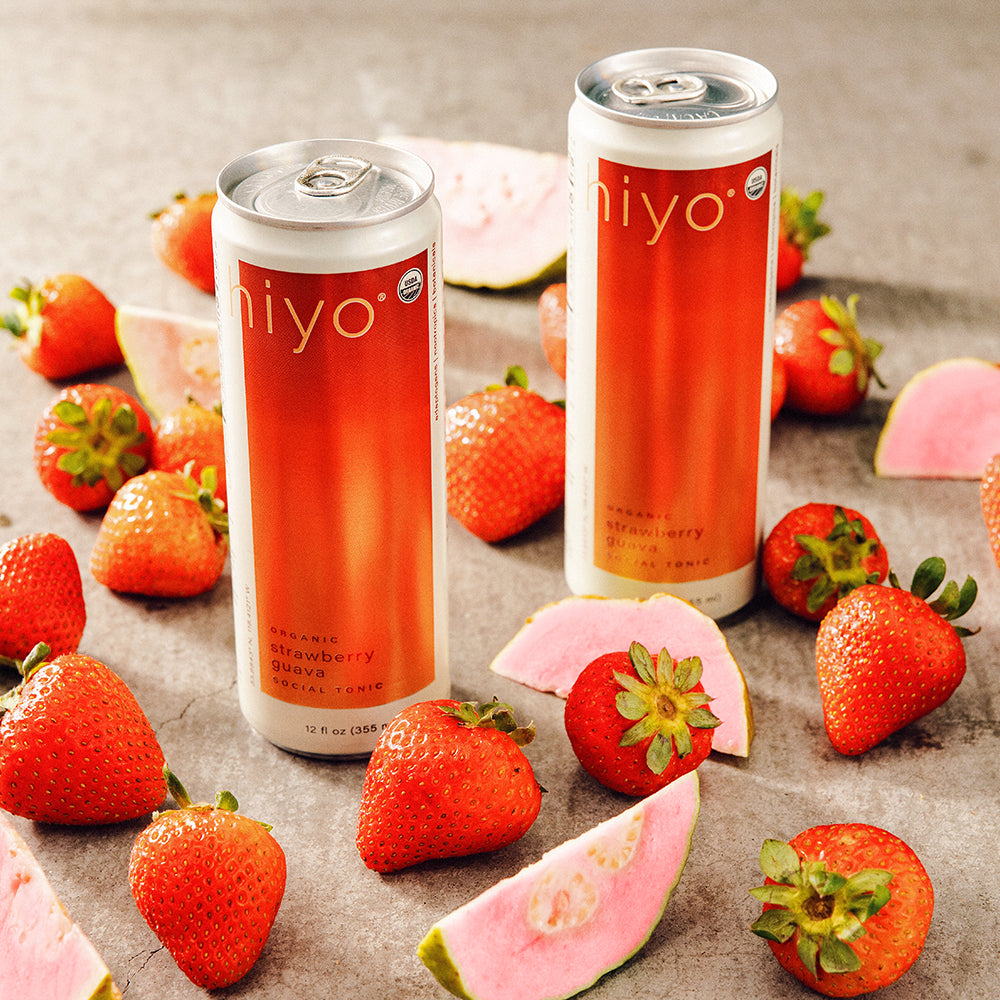 Two cans of hiyo strawberry drink surrounded by strawberries and guava slices on a textured surface.