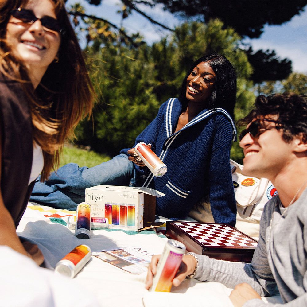 Three friends enjoying a picnic with colorful drinks and a checkered board game outdoors.