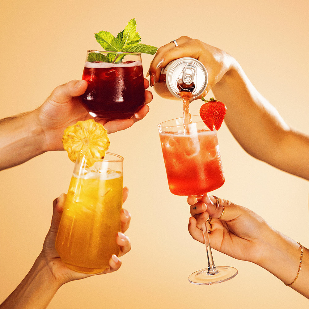 Four hands holding drinks with a can being poured into one of them against a beige background