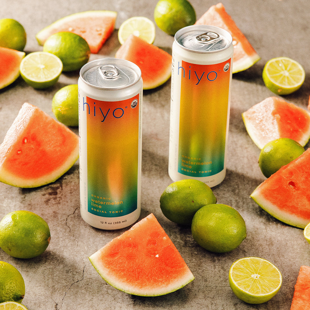 Two cans of hiyo watermelon lemon lime soda surrounded by watermelon slices and limes on a rustic surface.