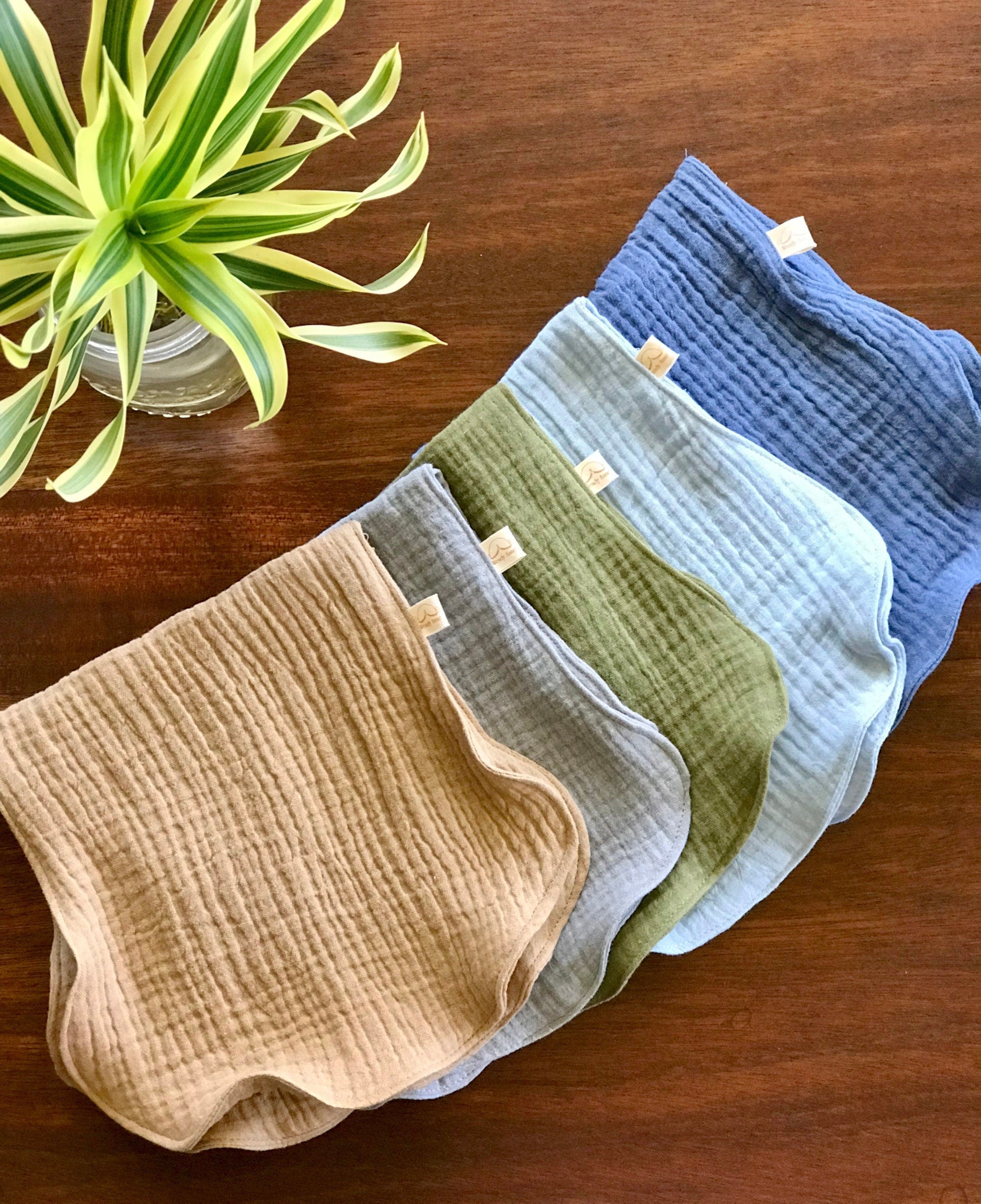 Set of five folded baby pants in beige, green, blue, and gray on a wooden surface with a plant in the corner.