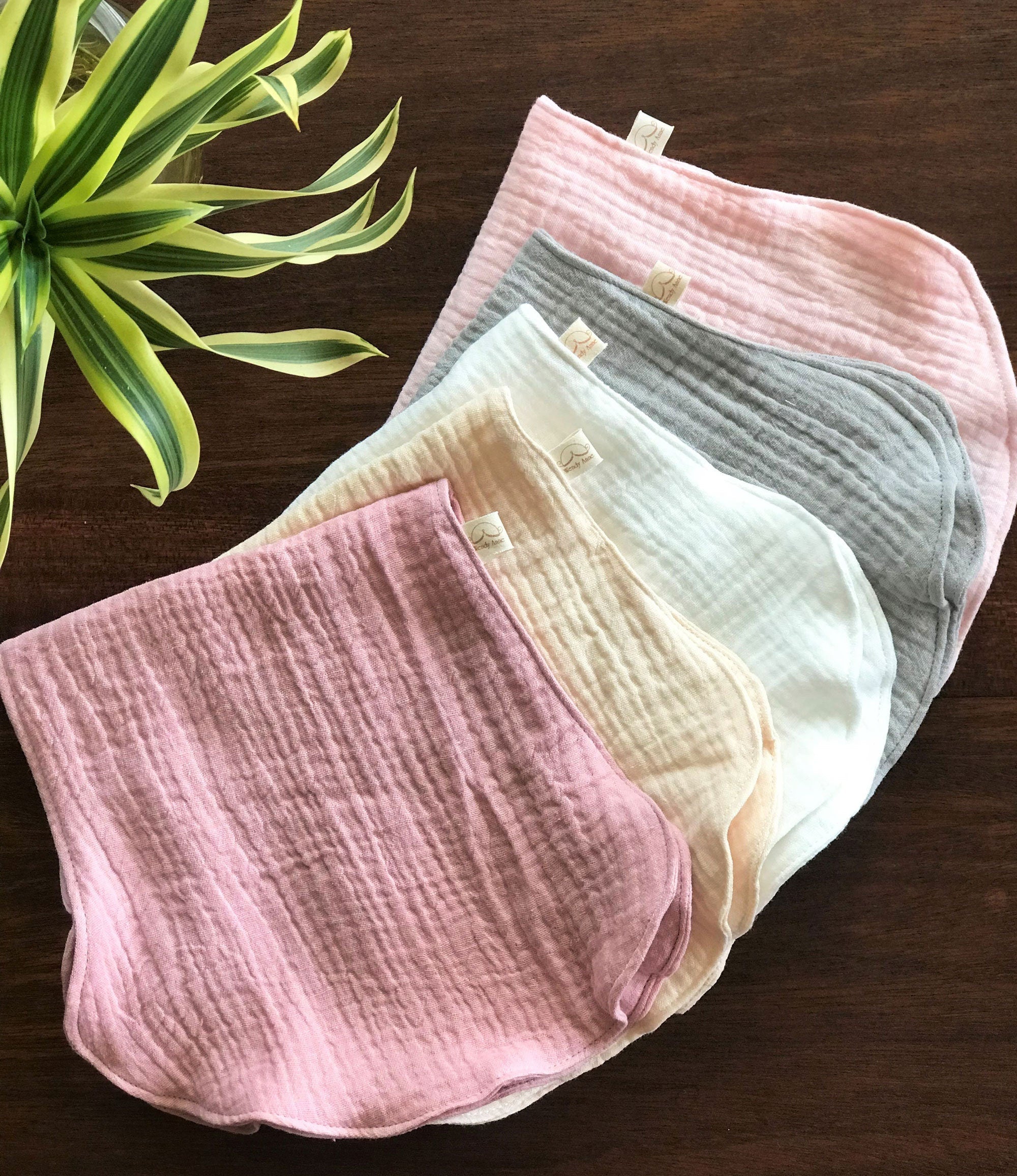 Set of pink, gray, and white baby bibs on a wooden surface with a plant in the corner.