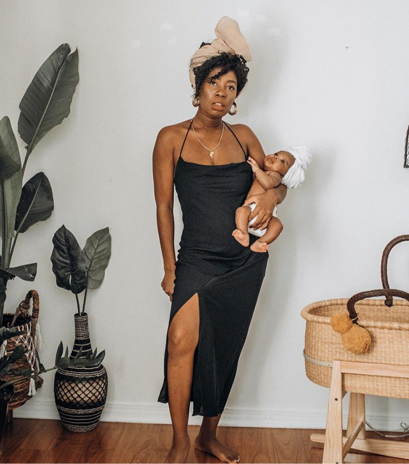 Woman in a black dress holding a baby in a home setting with plants and a basket.