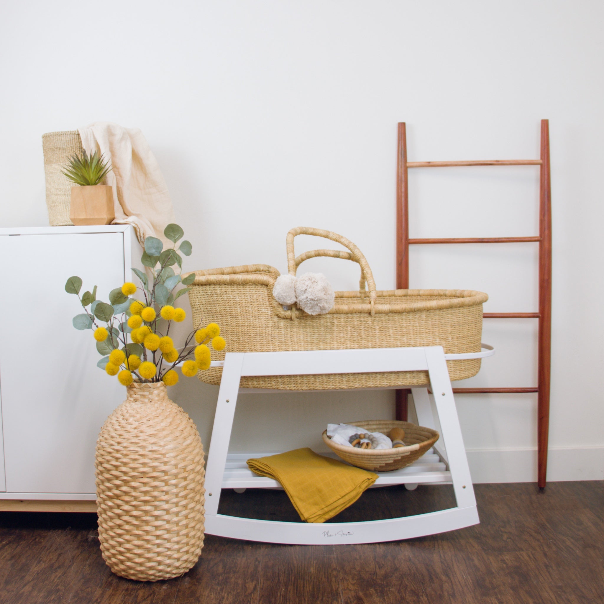 Nursery setup with wicker bassinet, white changing table, and decorative items on a wooden floor.