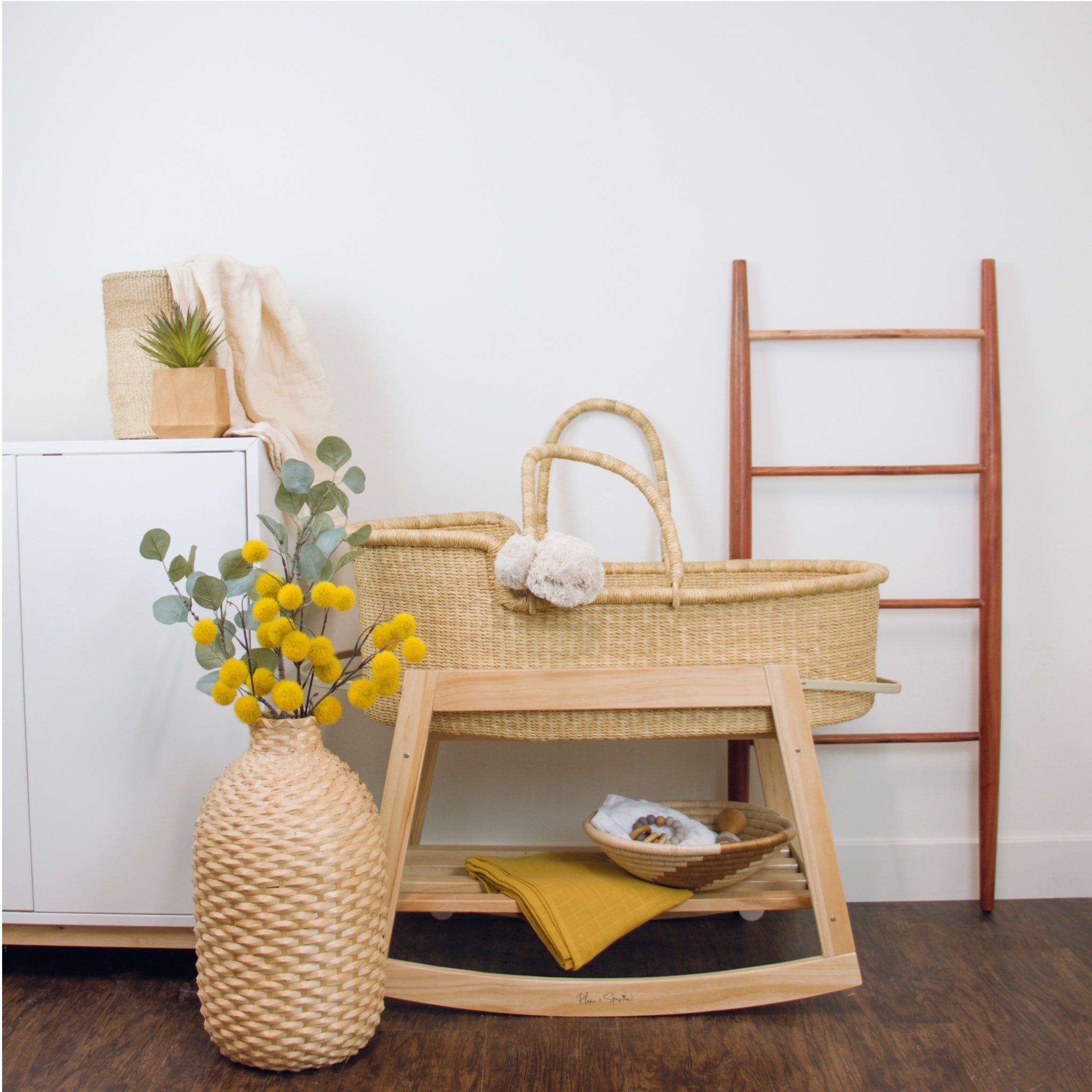 Wicker crib, vase with yellow flowers, and wooden ladder against a white wall.