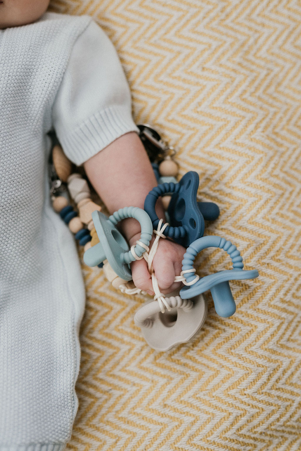 Child's hand holding a teething ring with wooden beads on a yellow and white patterned blanket.