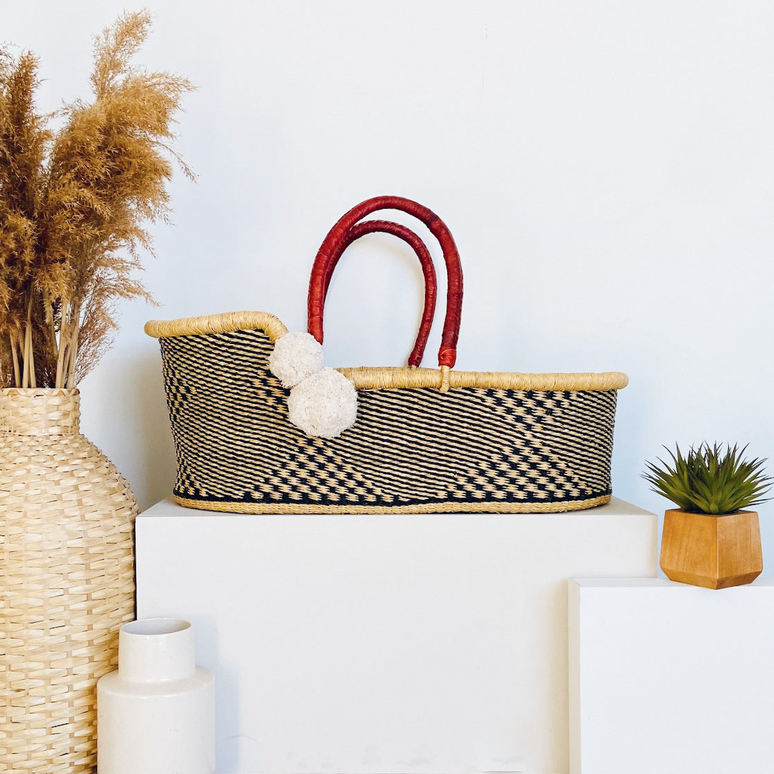 Woven basket with red handles on a white surface with decorative plants and vases.