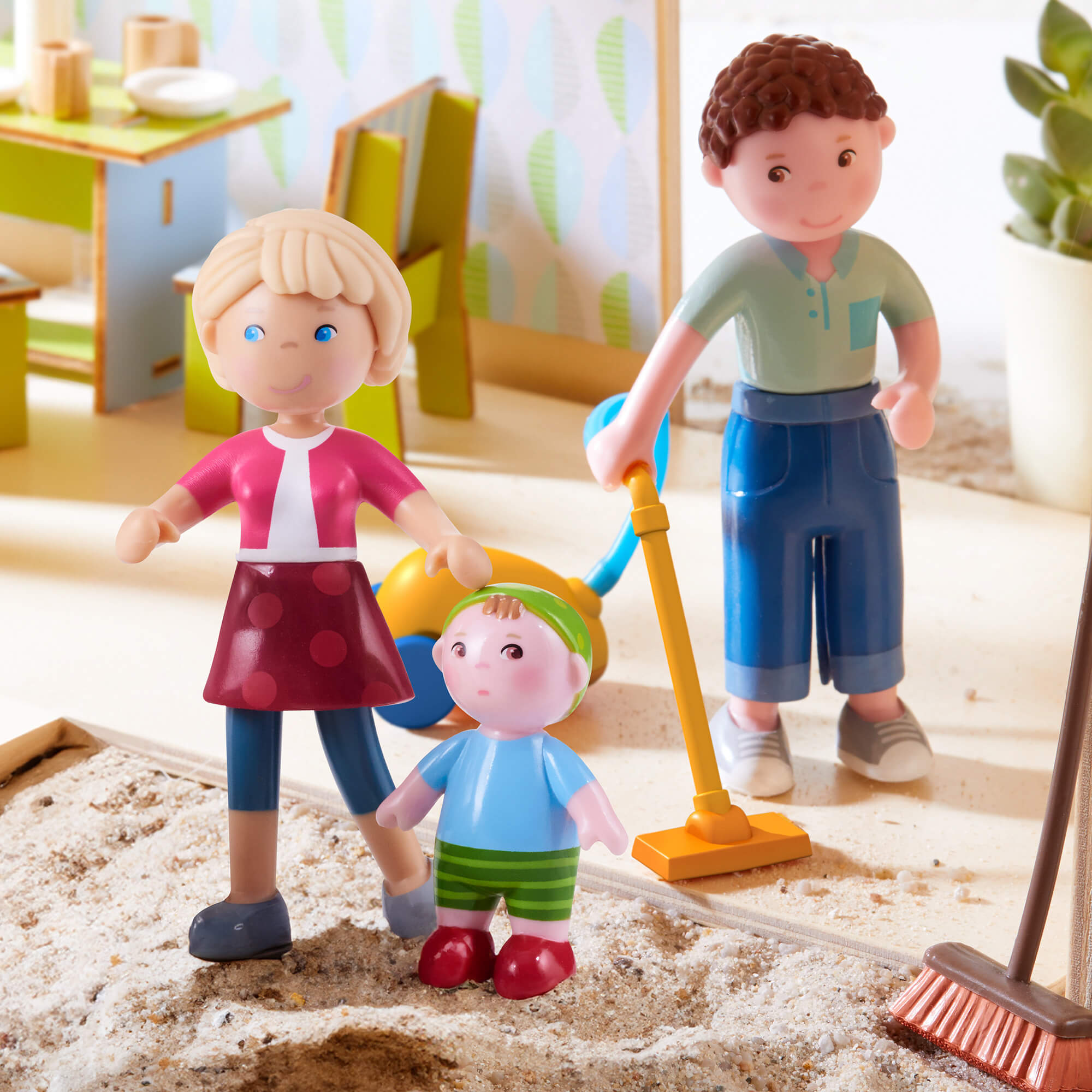 Toy figures of a family playing in a sand pit with a broom and toy shovel.