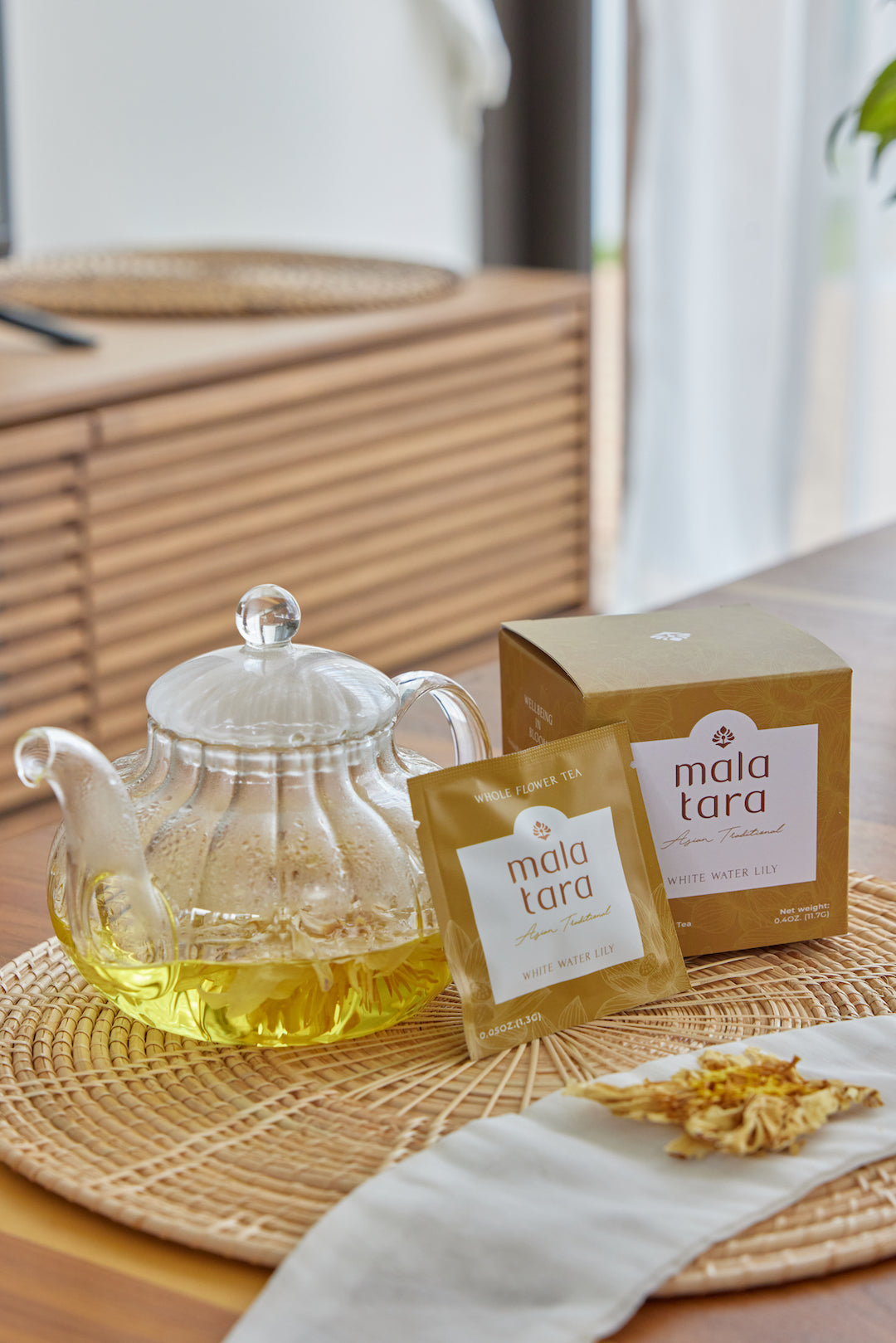 Glass teapot with yellow liquid, Mala Tara tea bags, and box on a woven mat.