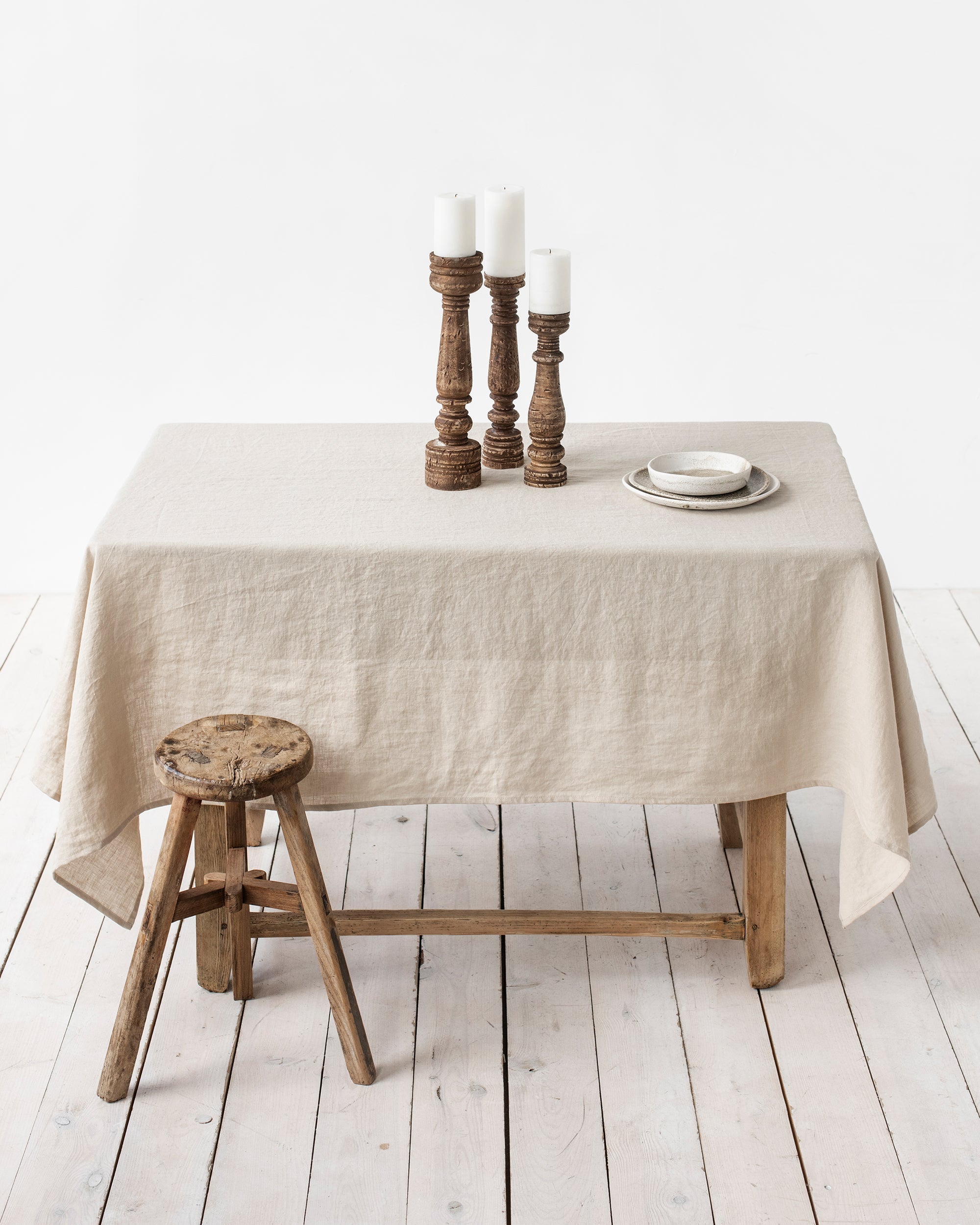 Beige tablecloth on a wooden table with candles and a stool in a minimalistic setting.