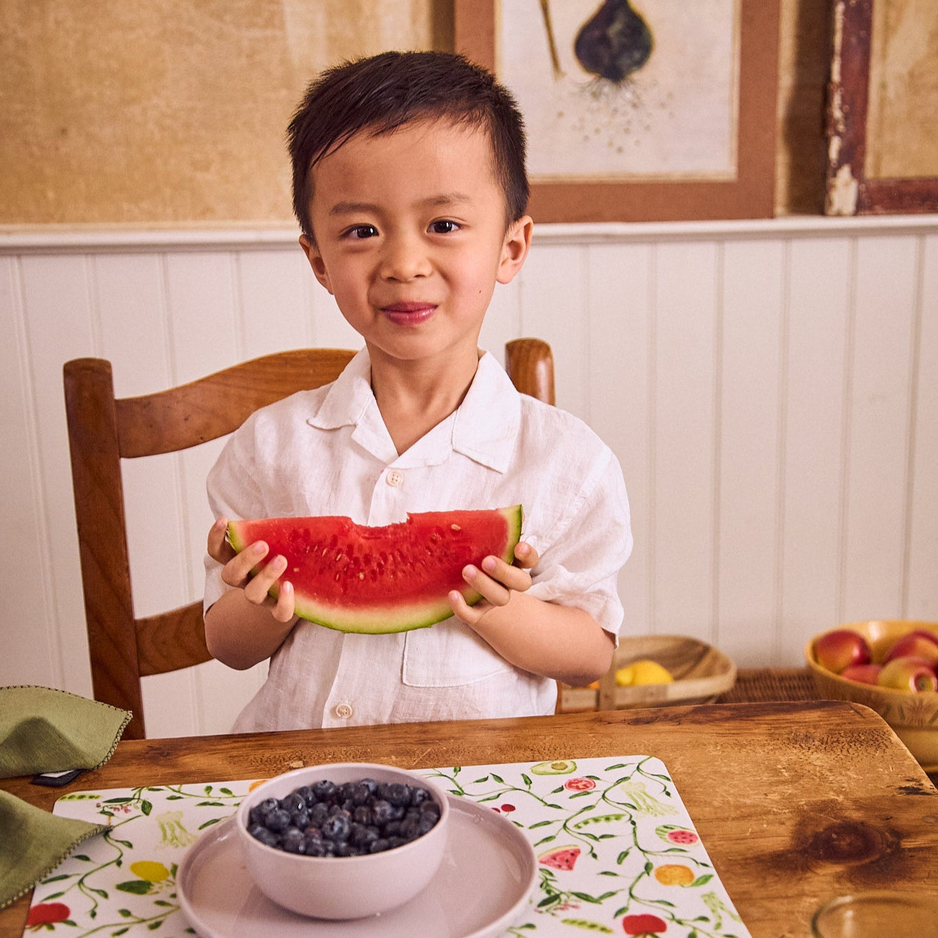 Child holding a slice of watermelon at a dining table with a bowl of blueberries.