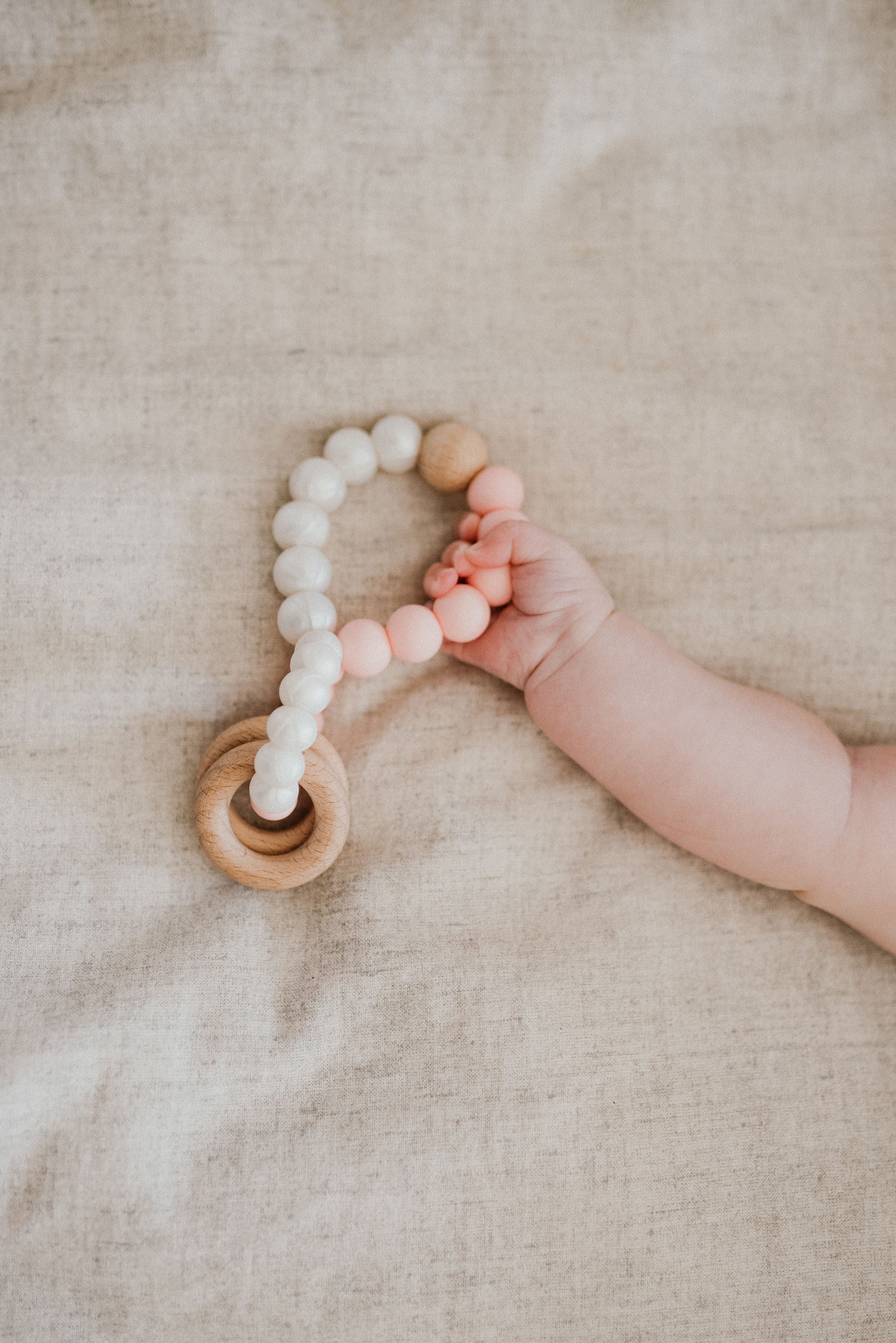 Baby hand holding a wooden teething ring with beads on a beige blanket
