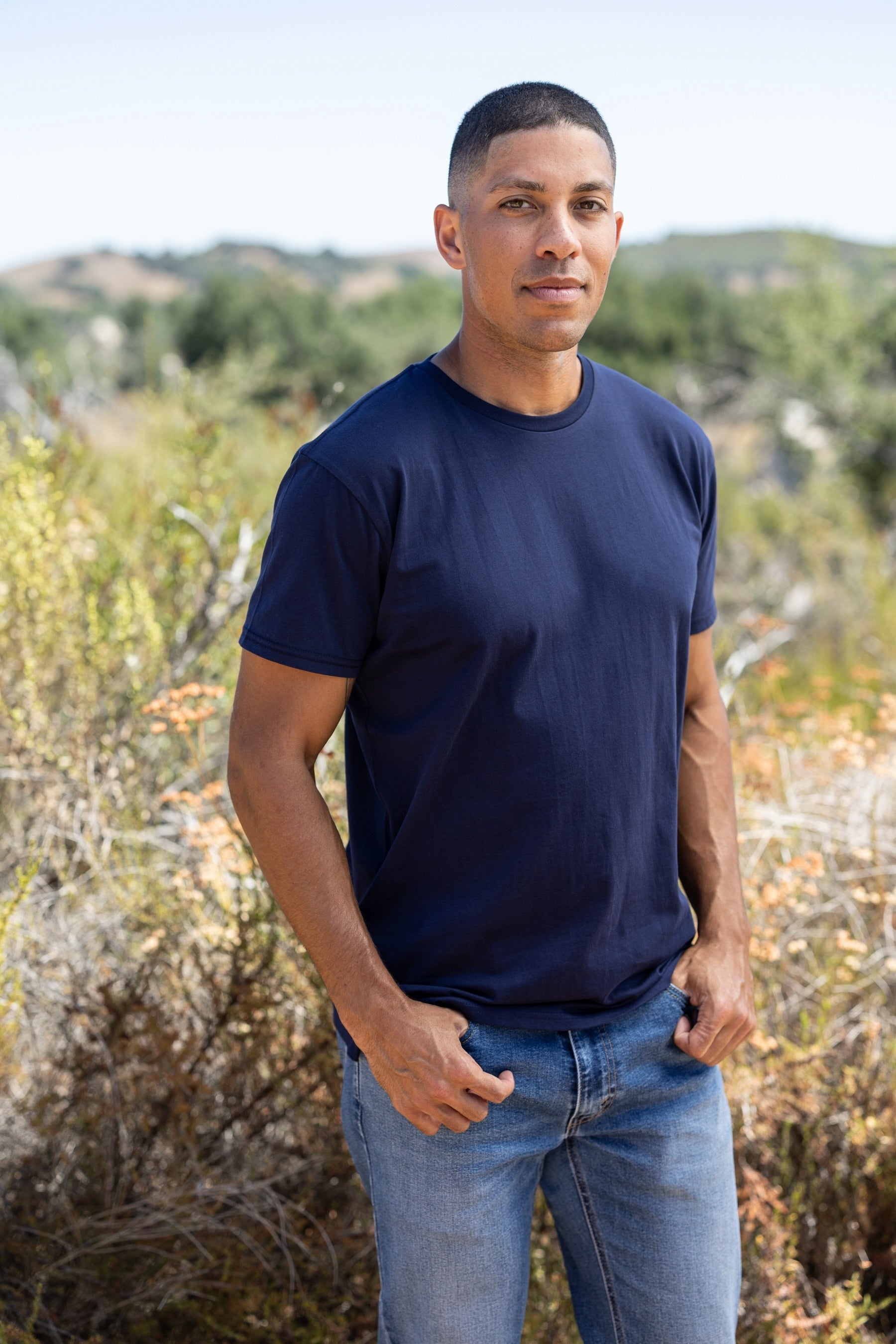 Man wearing a navy blue t-shirt and jeans standing outdoors with greenery in the background