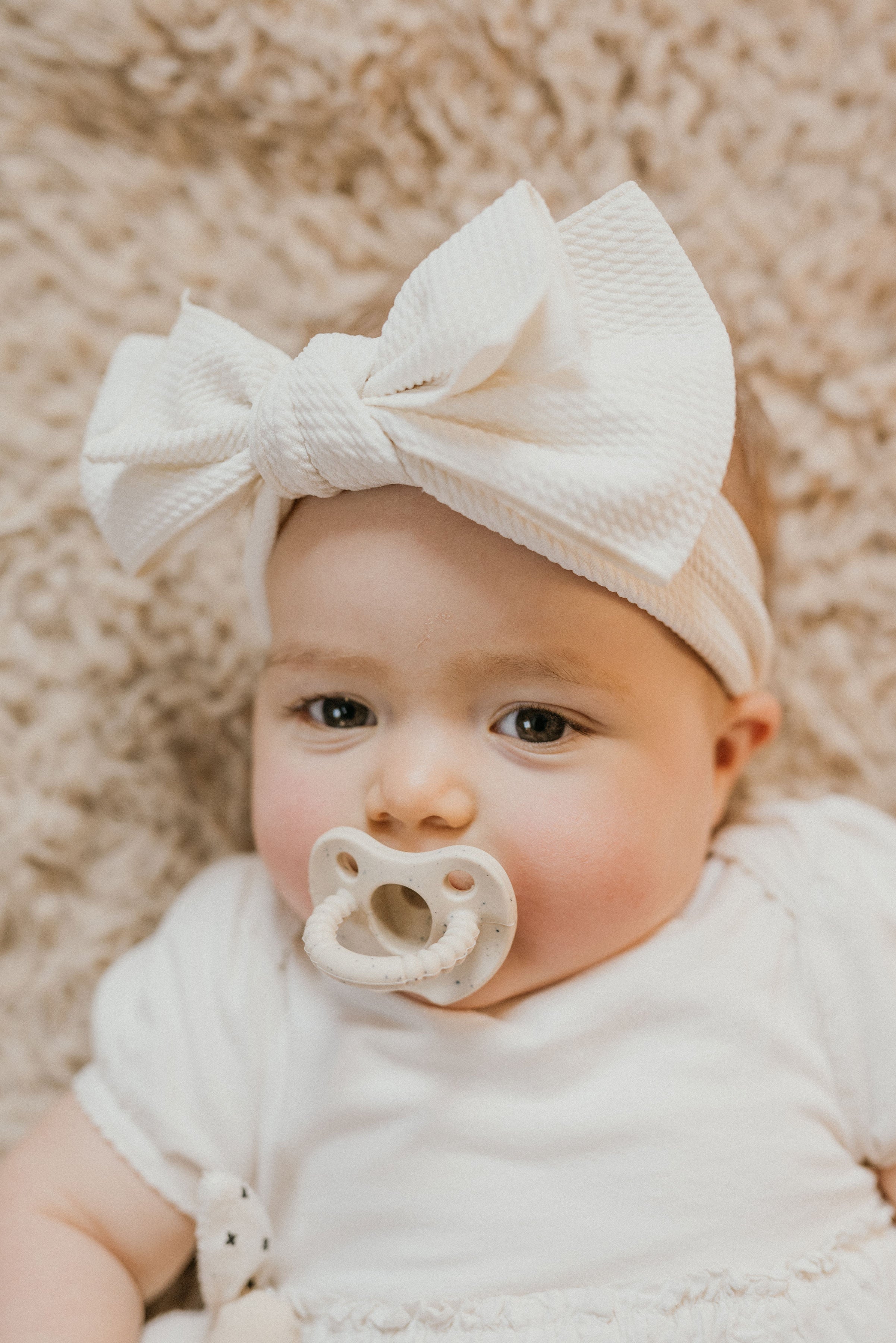 Baby wearing a white bow headband and pacifier on a soft beige background